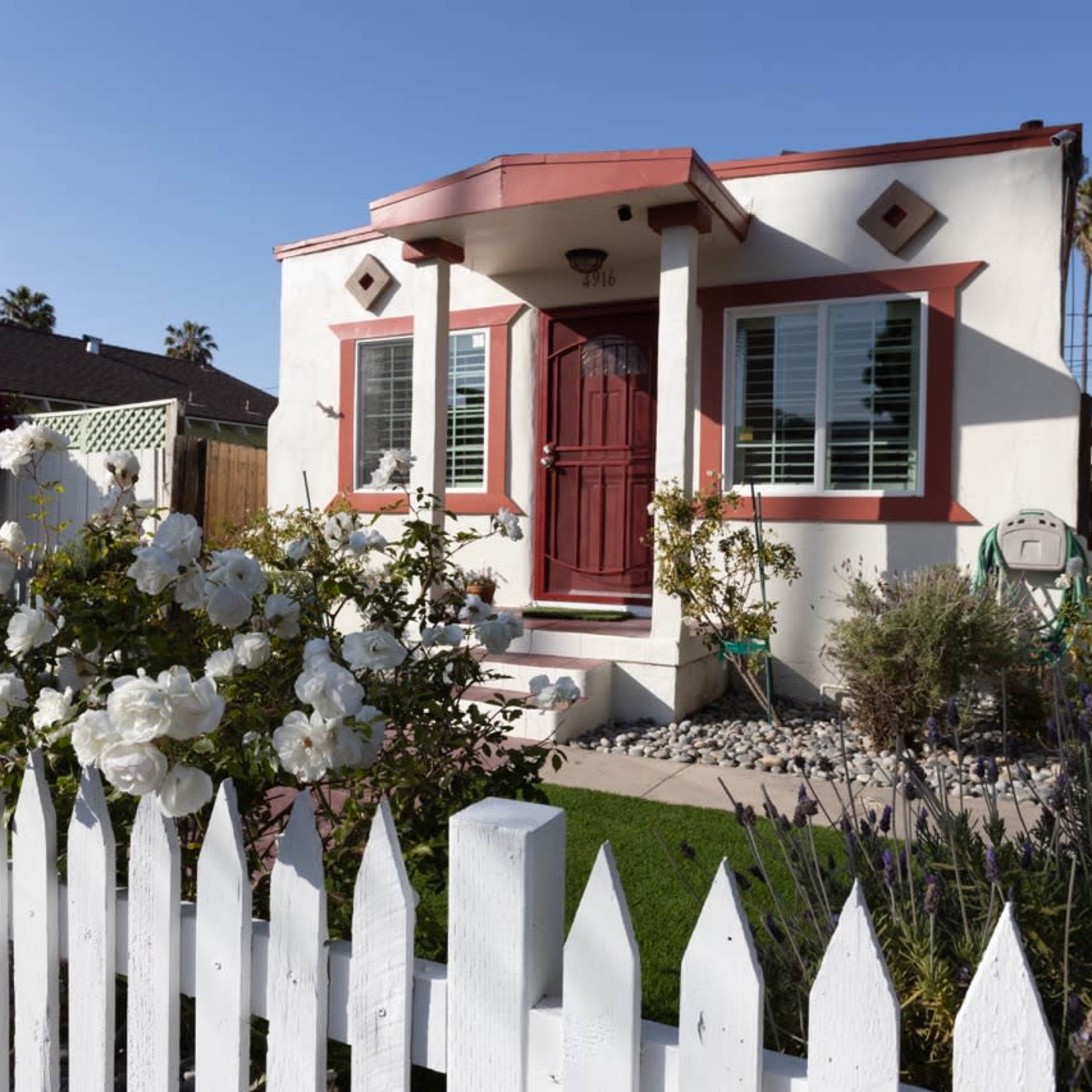 A small, white house with red trim features a front porch and a neatly kept garden surrounded by a white picket fence.
