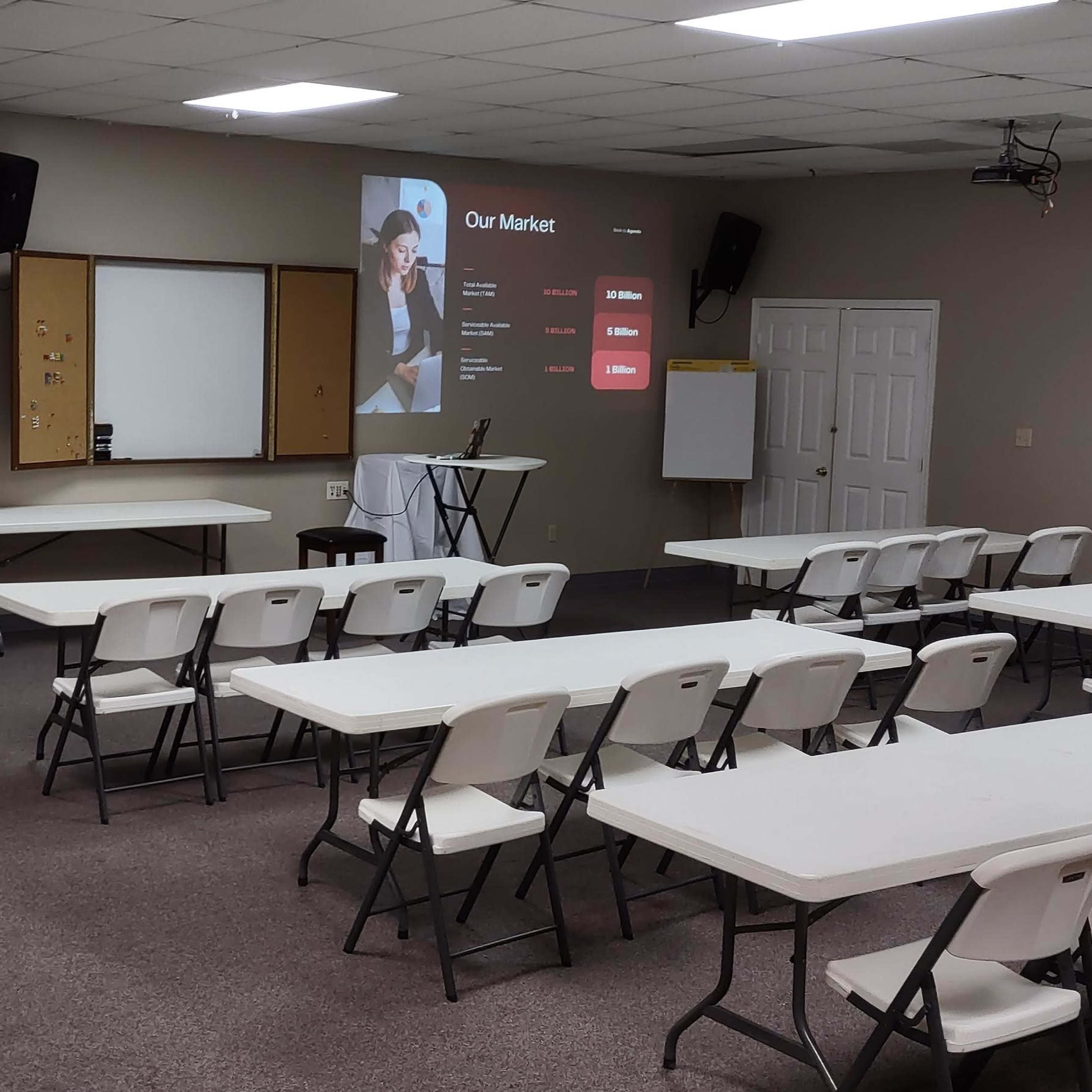 A classroom setting with rows of white tables and chairs, a projector screen displaying a presentation, and a black stand in the front.