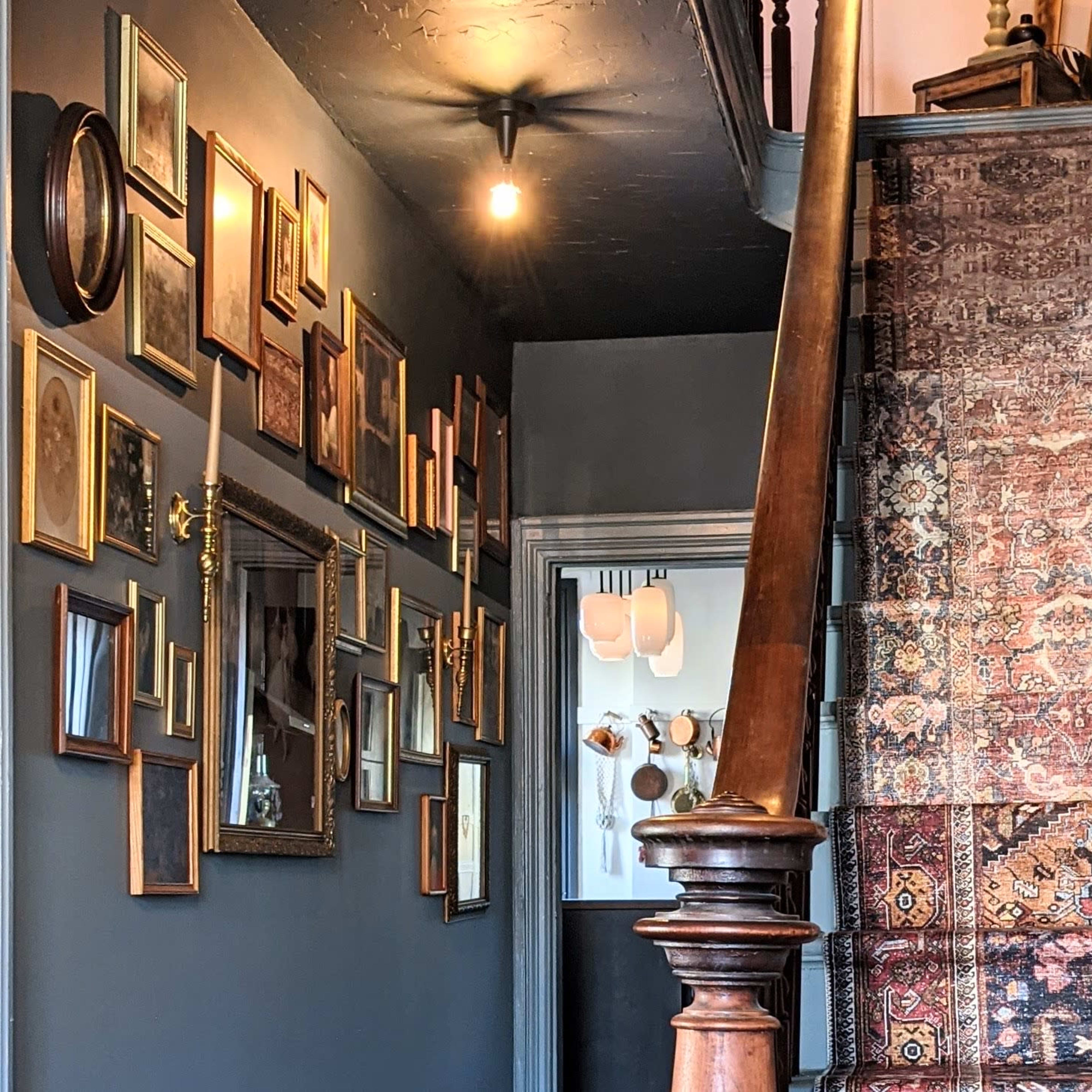 A staircase with a patterned rug, flanked by a wall decorated with various framed pictures, leading up to a dimly lit landing.