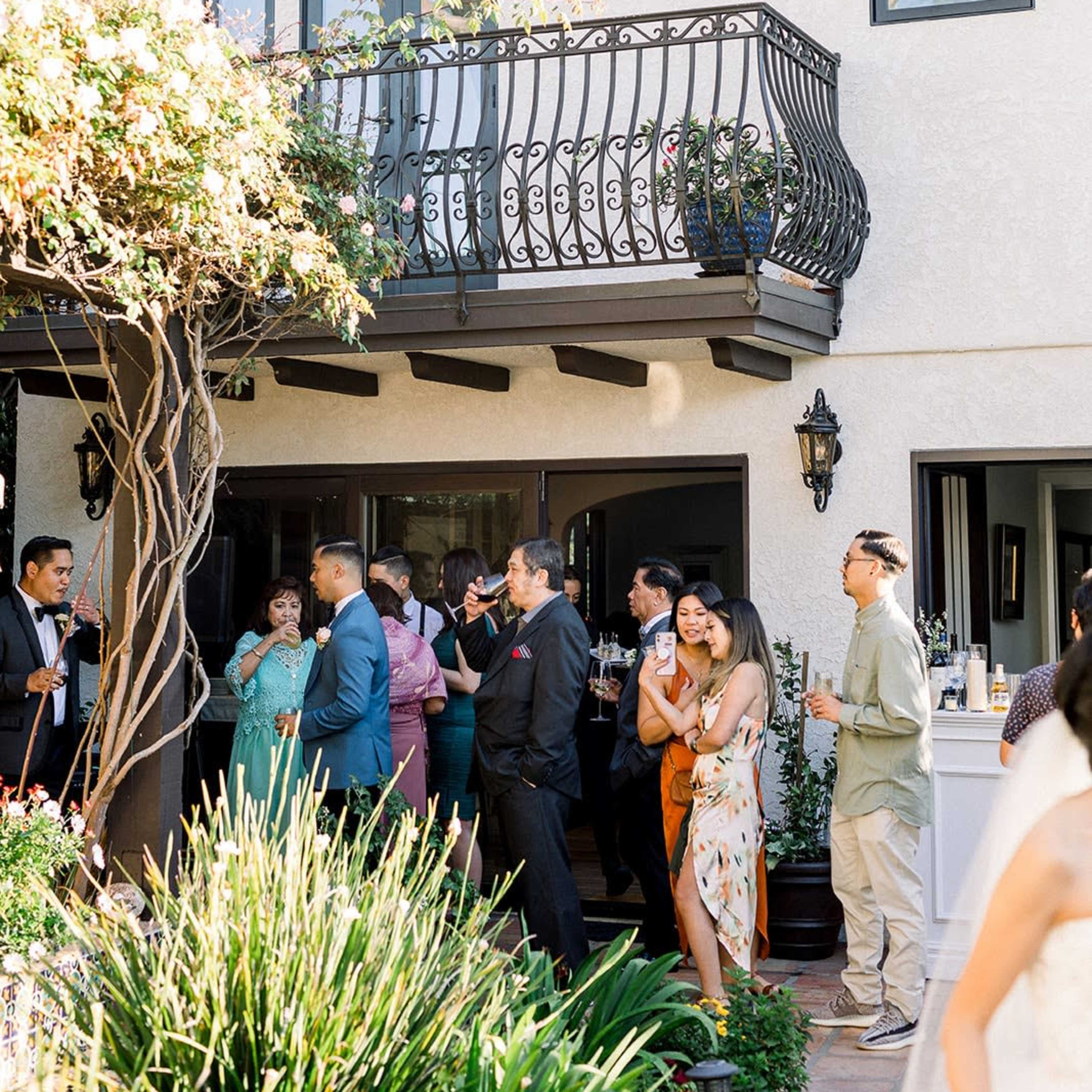 A group of people socializes outside a house with a balcony, surrounded by greenery and architectural details.