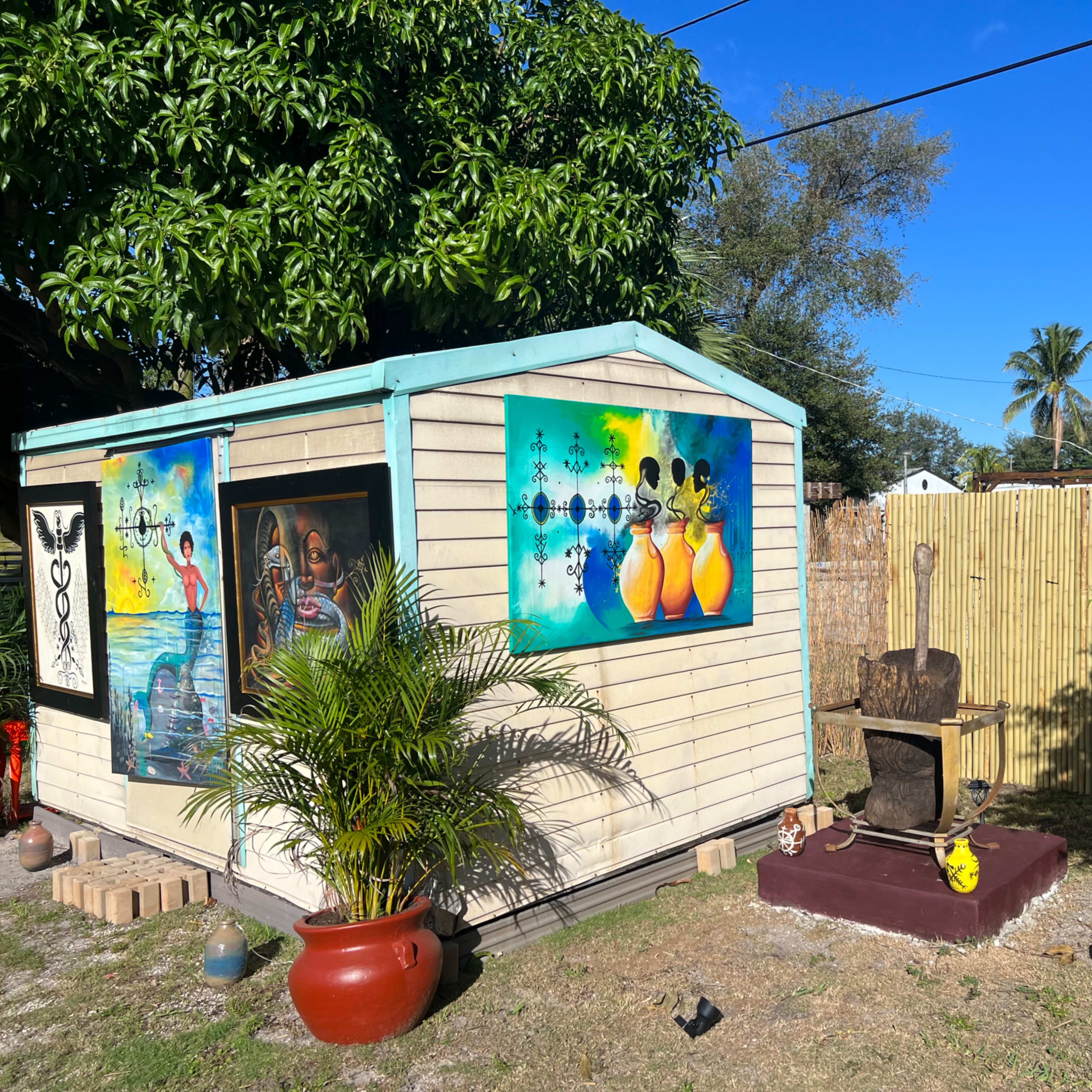 A small wooden shed with colorful artwork on the walls is surrounded by plants and decorative items in a yard.