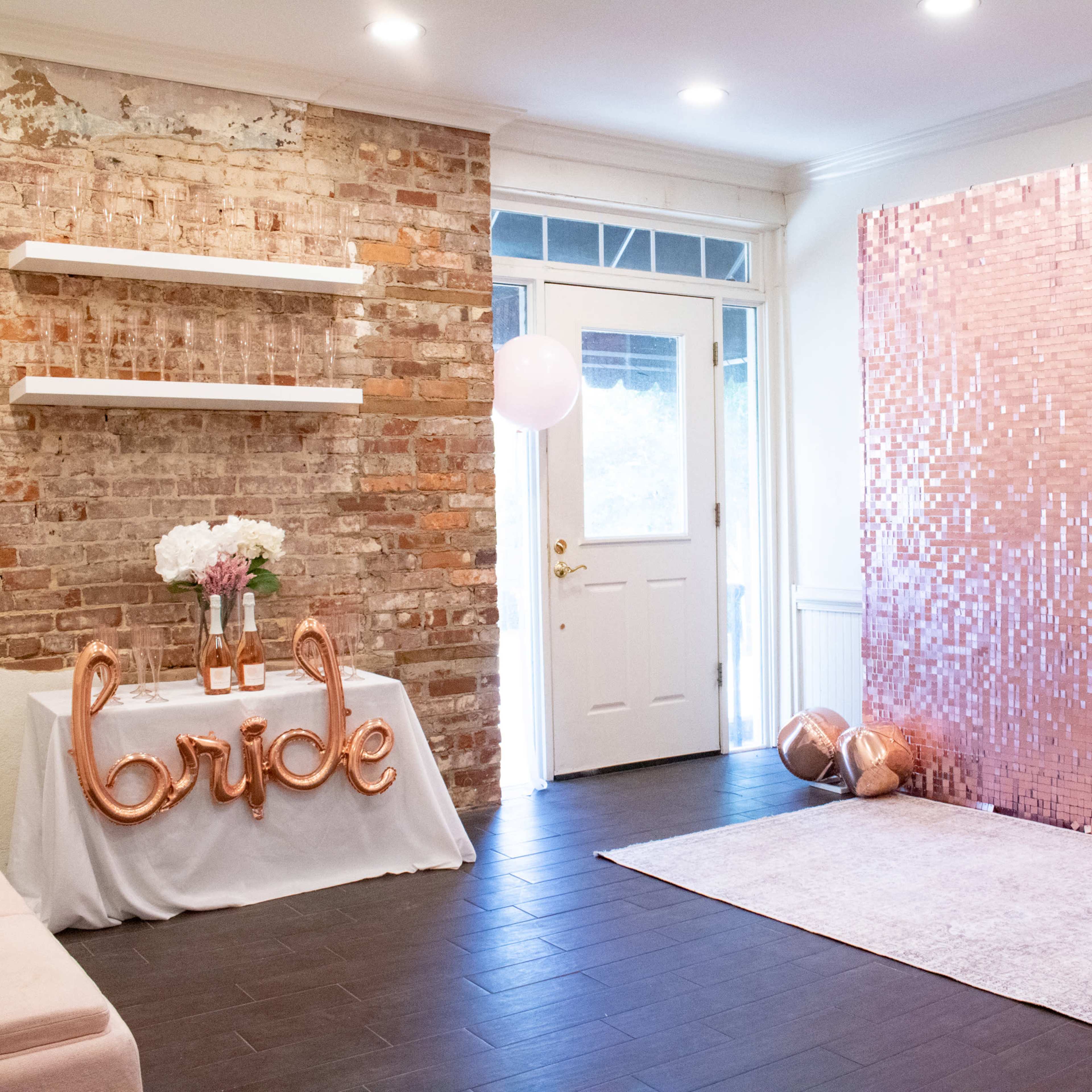 A decorated indoor space with a brick wall, a table displaying the word "bride" in metallic letters, a rose gold sequin backdrop, and decorative balloons.