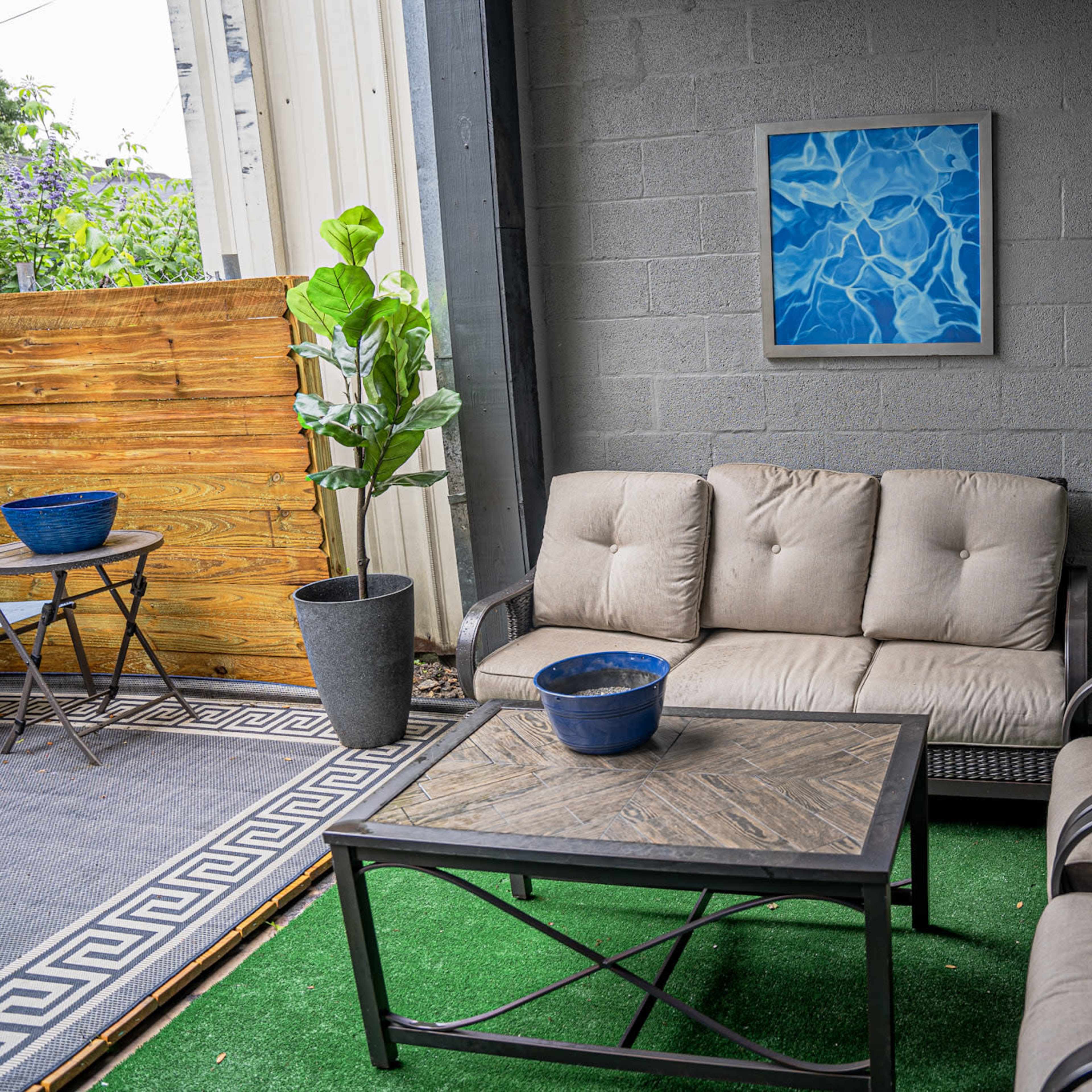 A cozy outdoor seating area with a beige couch, two armchairs, a coffee table, and decorative plants against a gray wall, featuring two framed blue abstract art pieces.