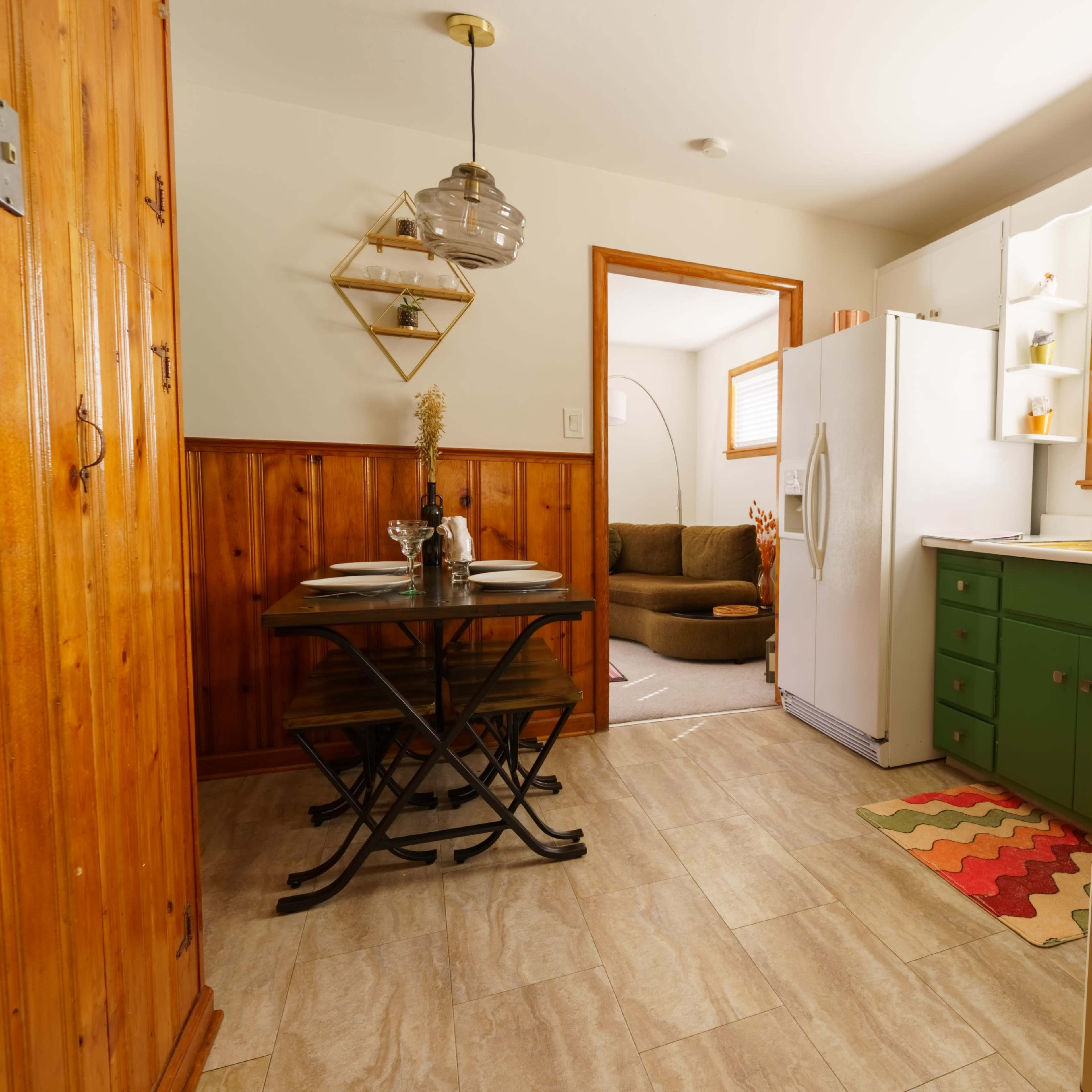 A cozy kitchen with green cabinets, a white refrigerator, a dining table set for two, and a comfortable living room visible in the background.