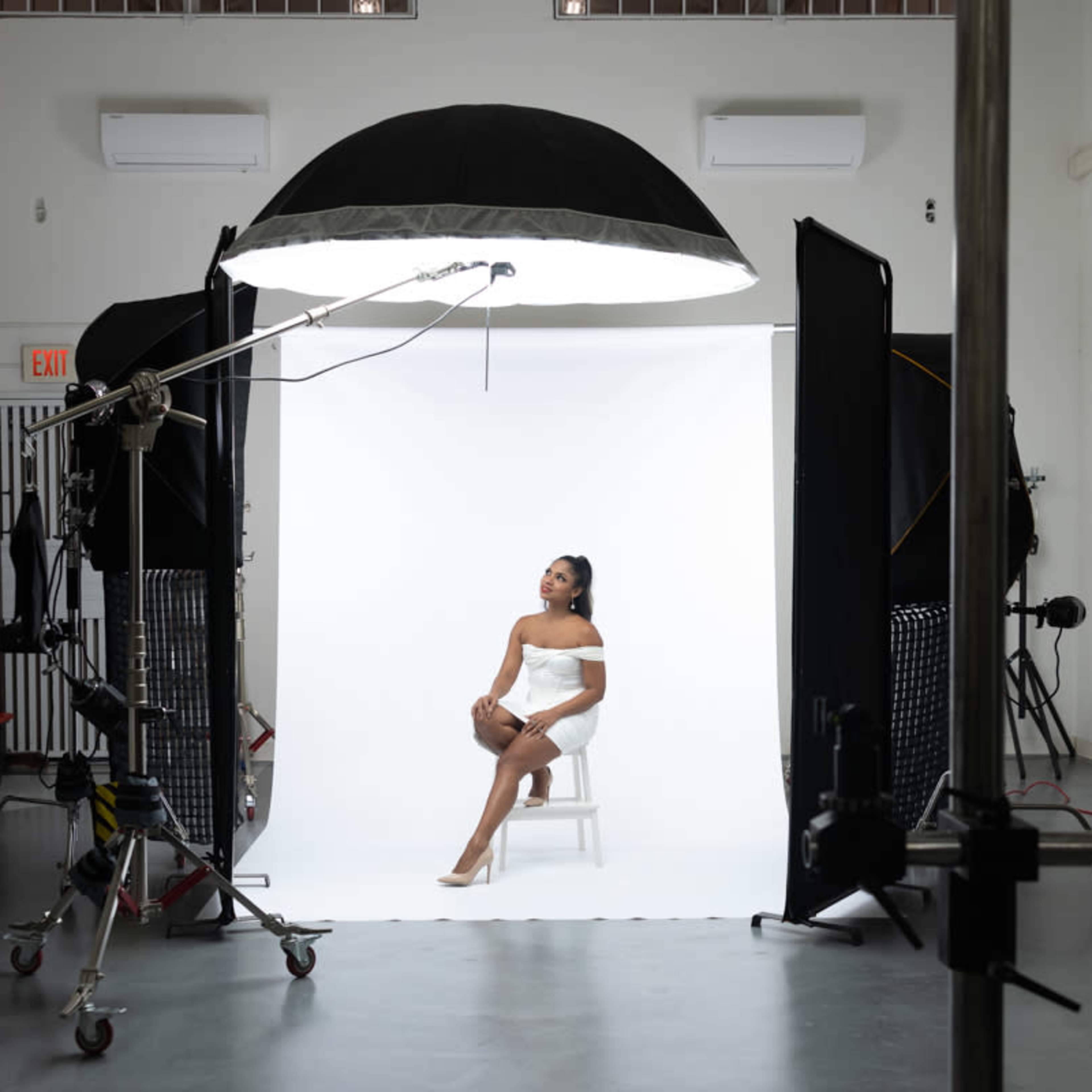 A model poses on a chair against a white backdrop in a photography studio equipped with various lighting and support equipment.