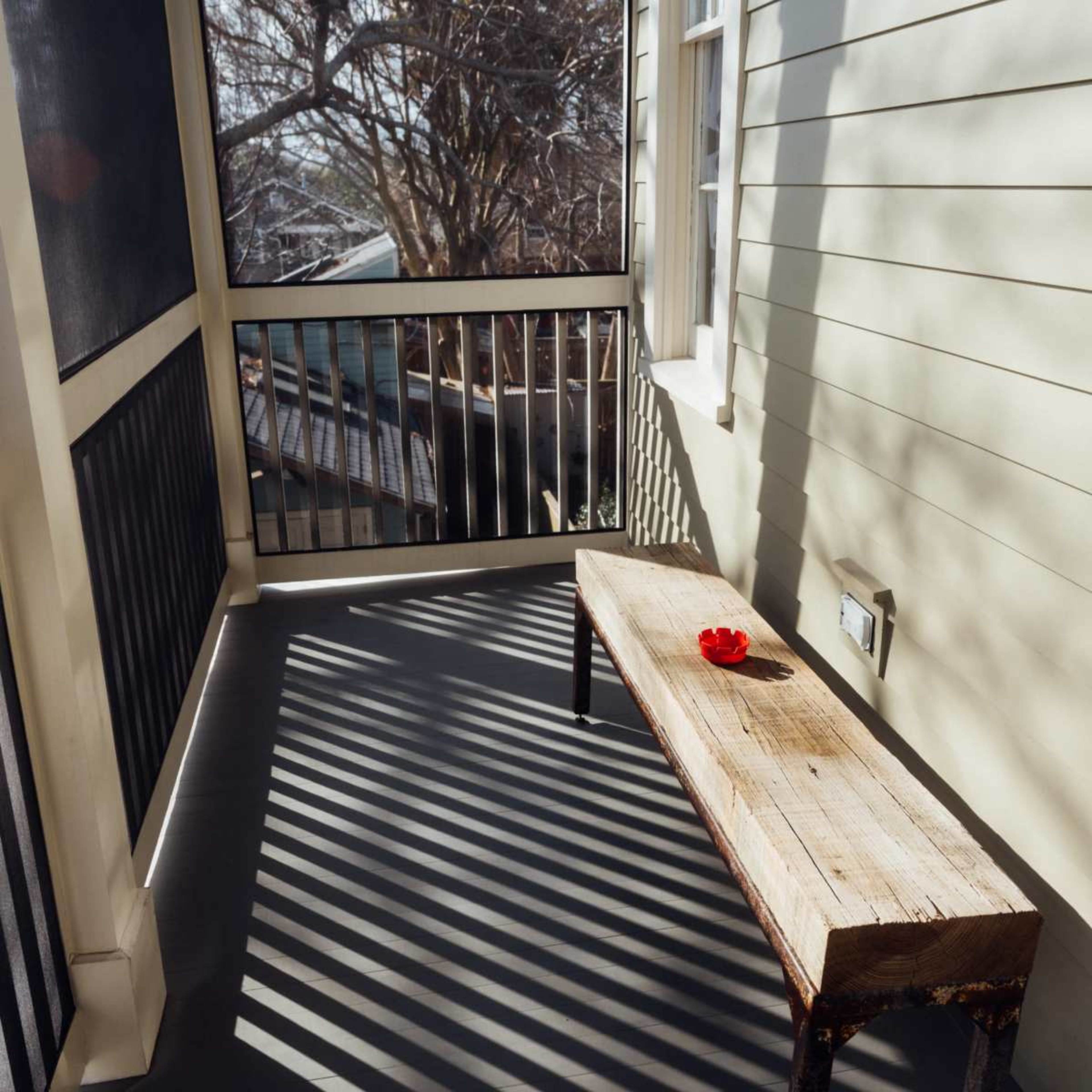 A narrow porch with a wooden bench and a red flower on it, surrounded by vertical shadows cast by nearby railings.