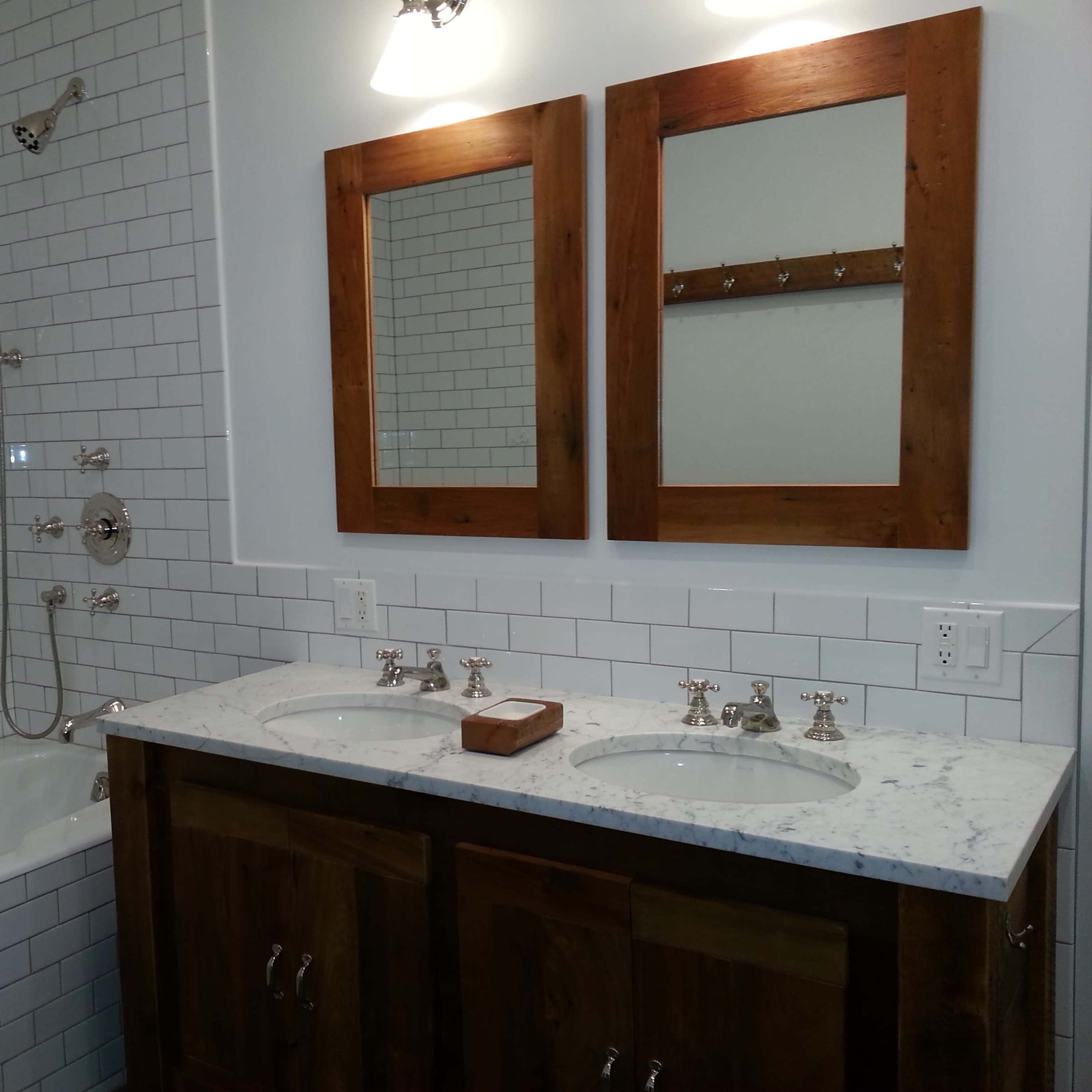 A bathroom featuring a white subway tile shower and bathtub, dual sinks with a marble countertop, and two wooden-framed mirrors hanging above.