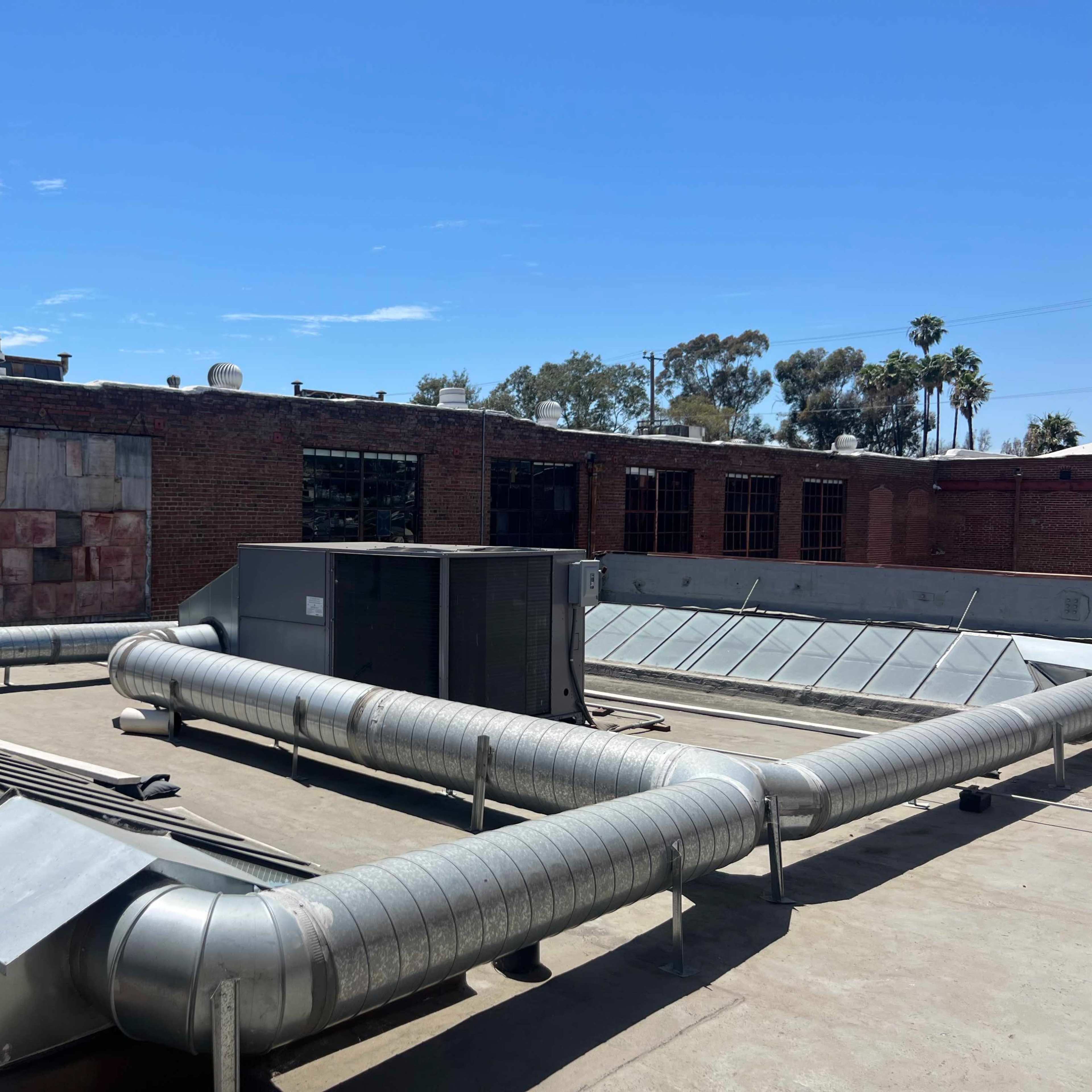 A rooftop with metal ductwork and an air conditioning unit, alongside a brick building with several windows.