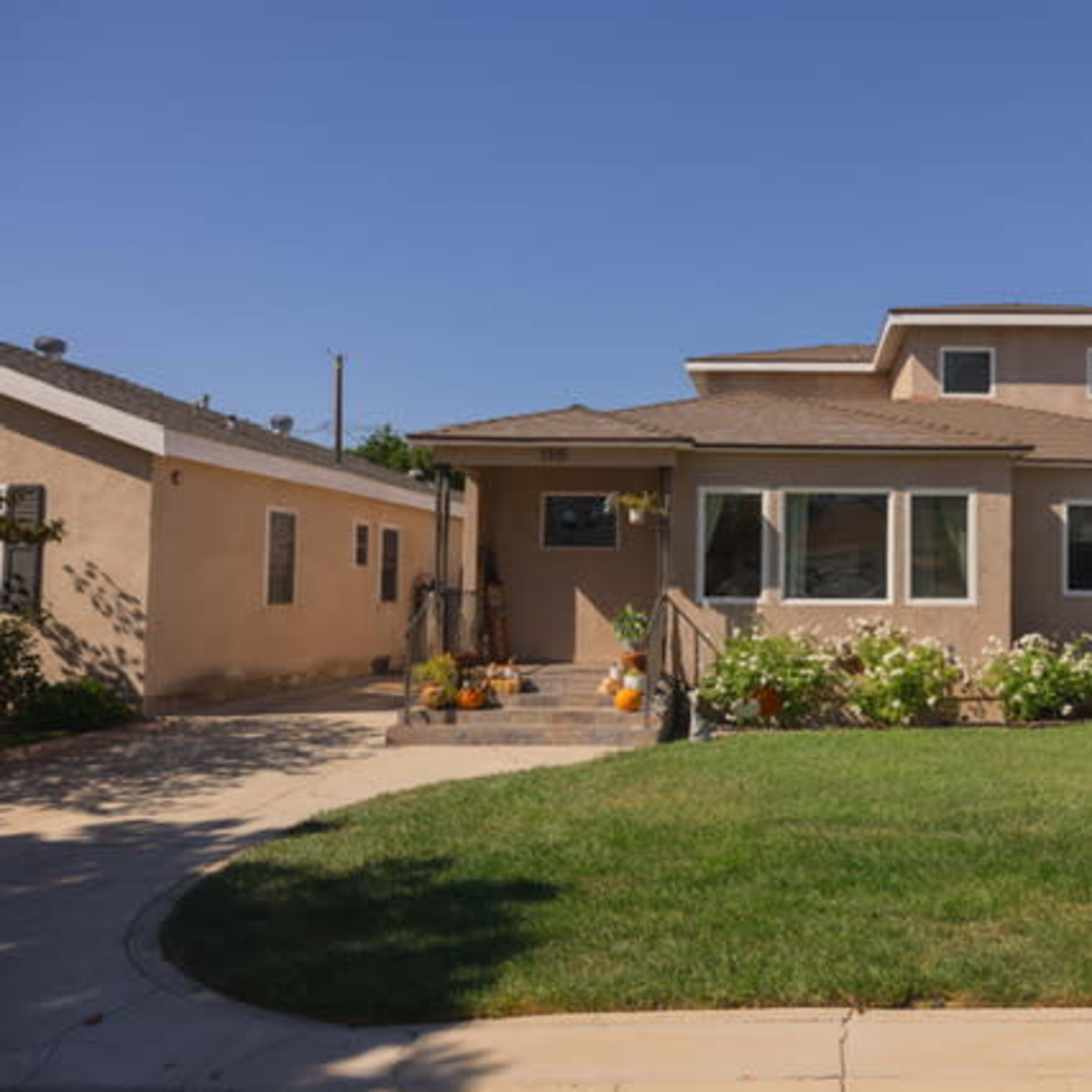 A two-story house with a landscaped front yard, featuring a mix of concrete driveway and light-colored exterior walls.