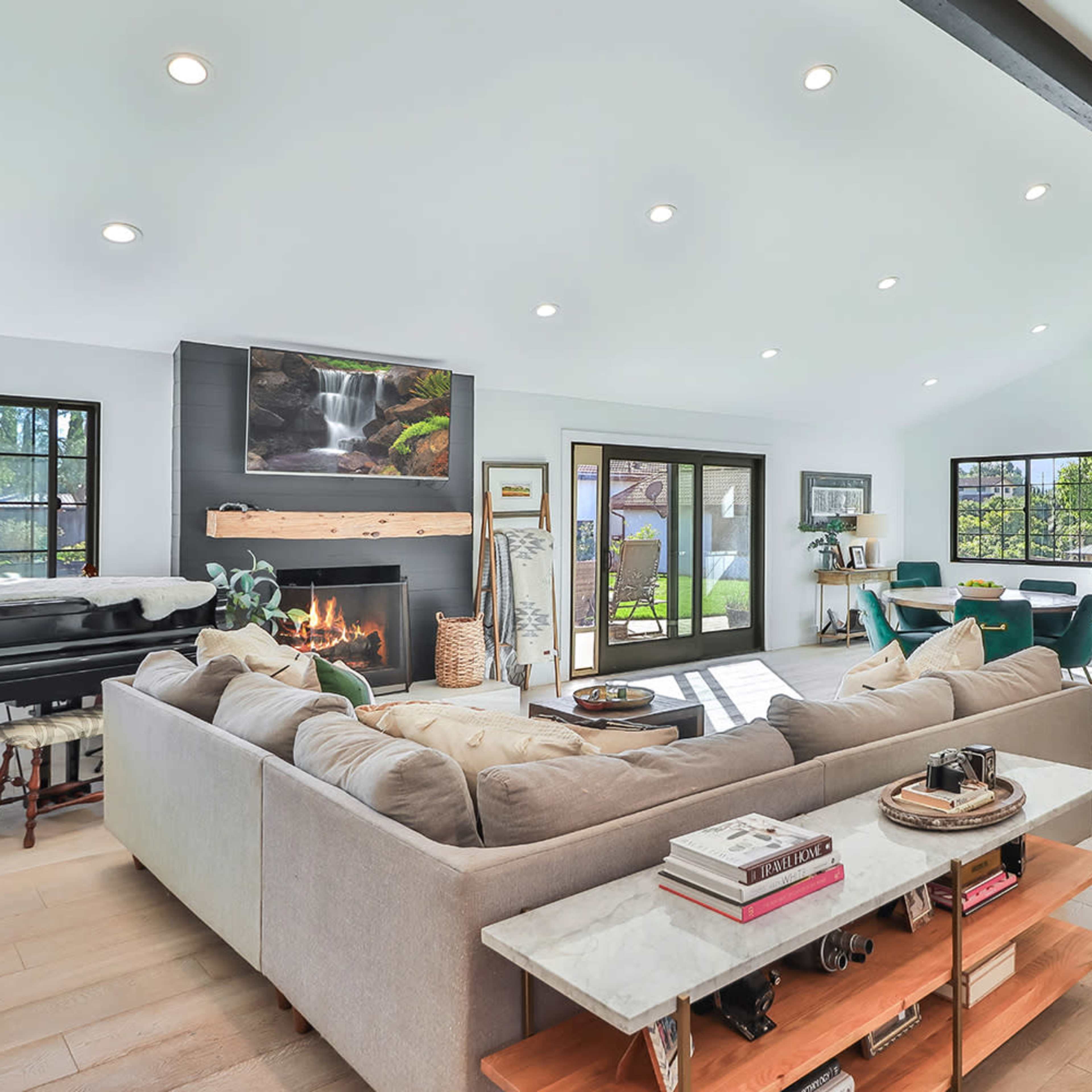 A modern living room with a sectional sofa, a fireplace, a piano, and large windows overlooking a backyard.