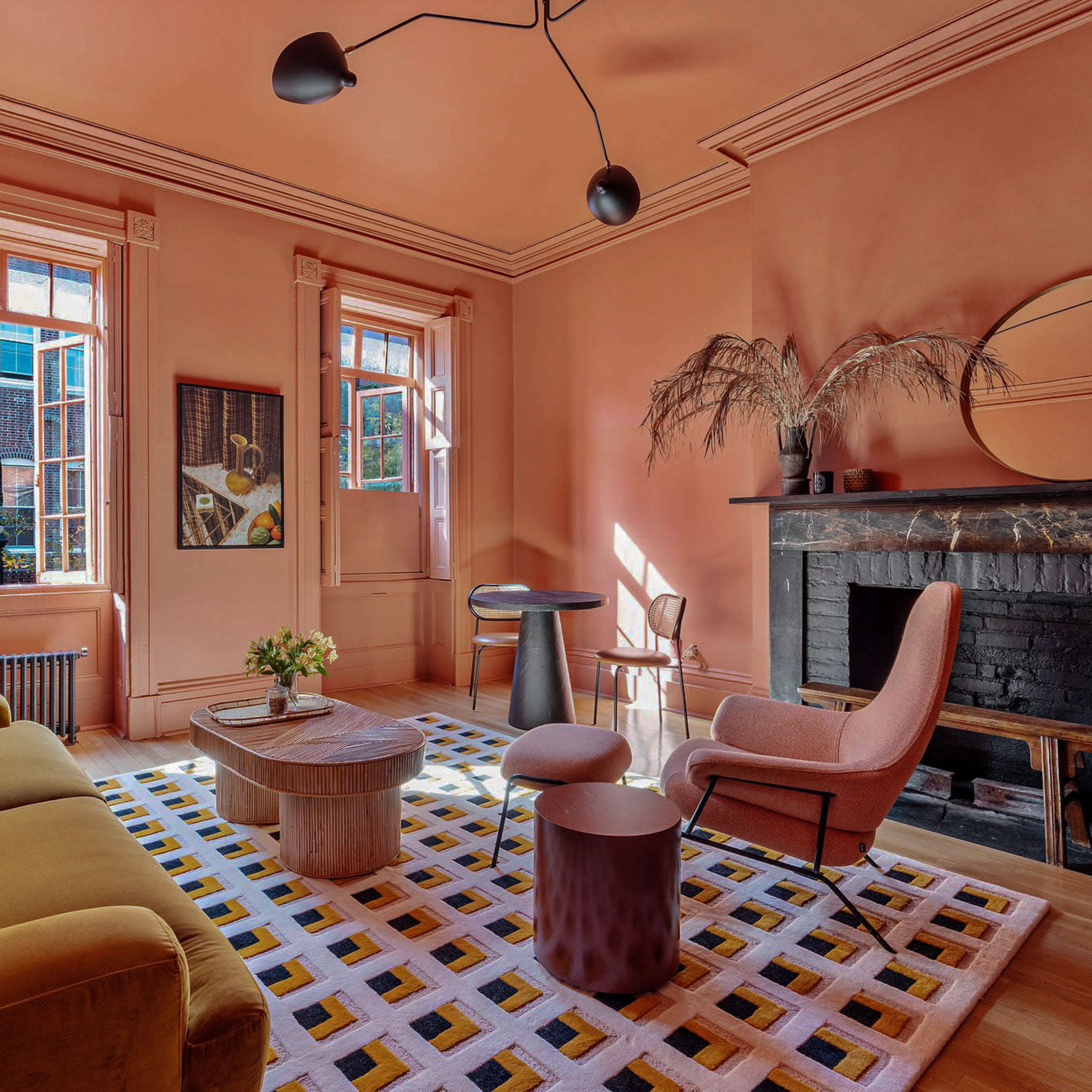 The room features a pink-walled living area with a yellow sofa, a round wooden coffee table, an armchair, and a black fireplace, illuminated by natural light through large windows.