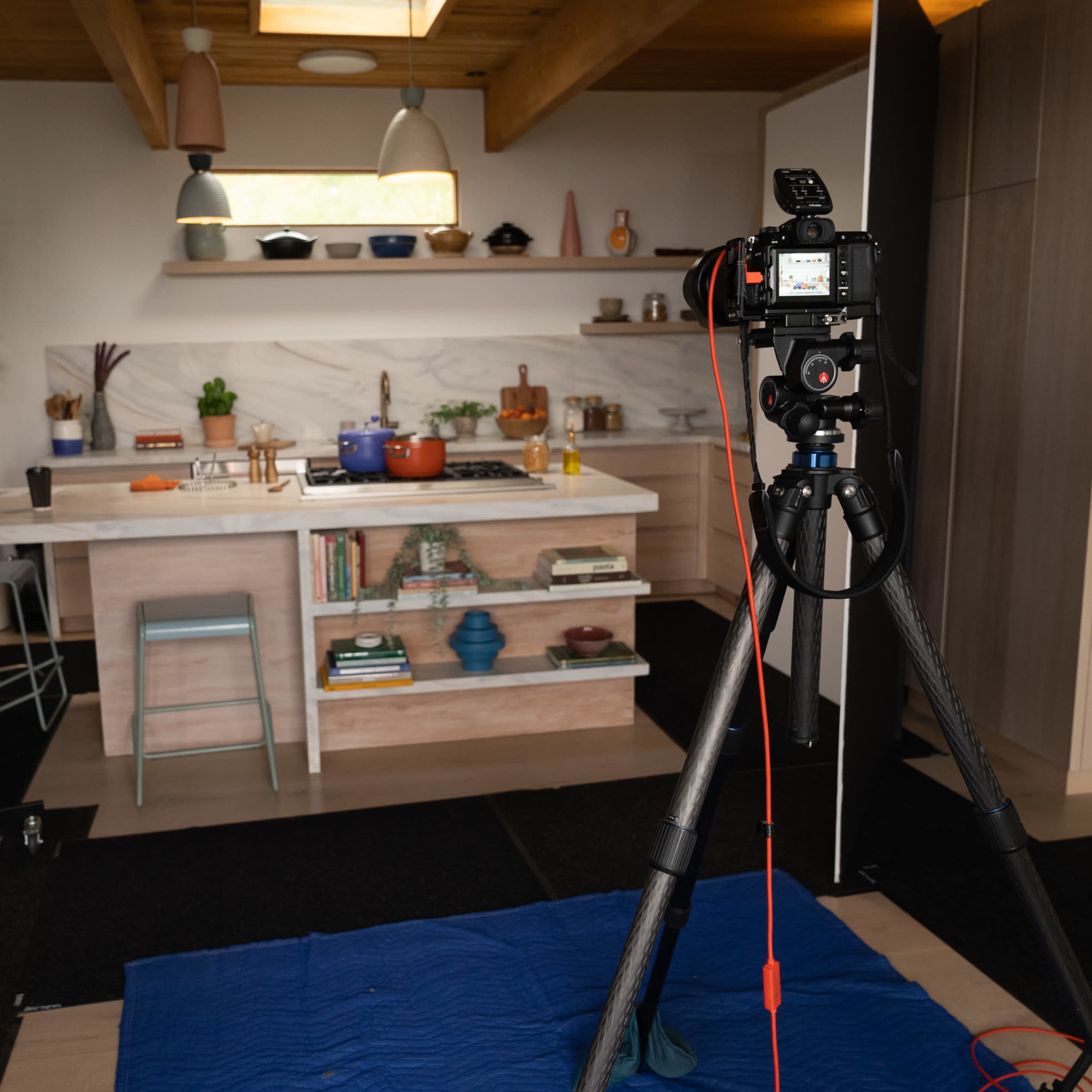 A photography setup is arranged in a kitchen, featuring a camera on a tripod facing a marble countertop with various cooking utensils and plants in the background.