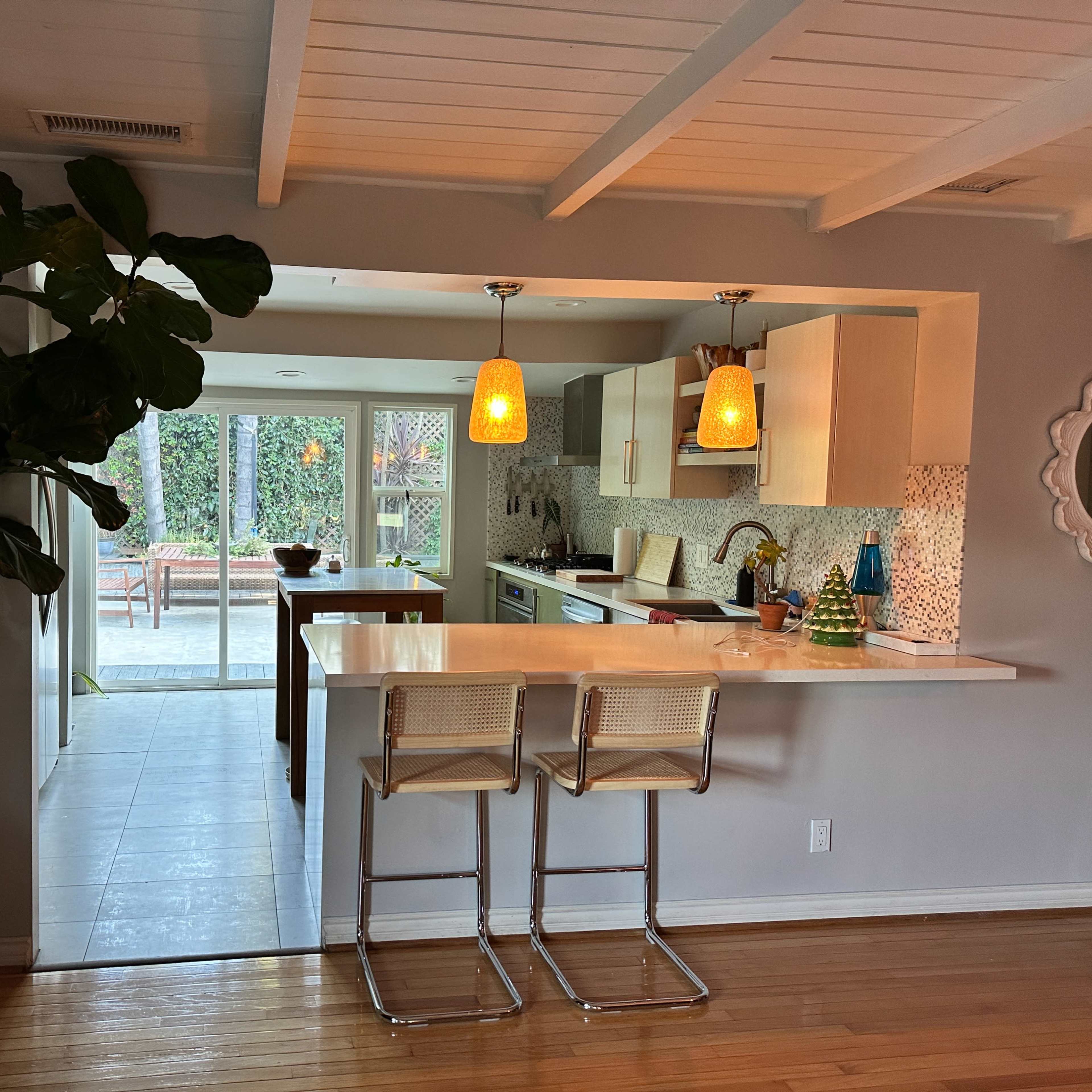 A modern kitchen with two bar stools, pendant lighting, and a large window leading to an outdoor patio area.