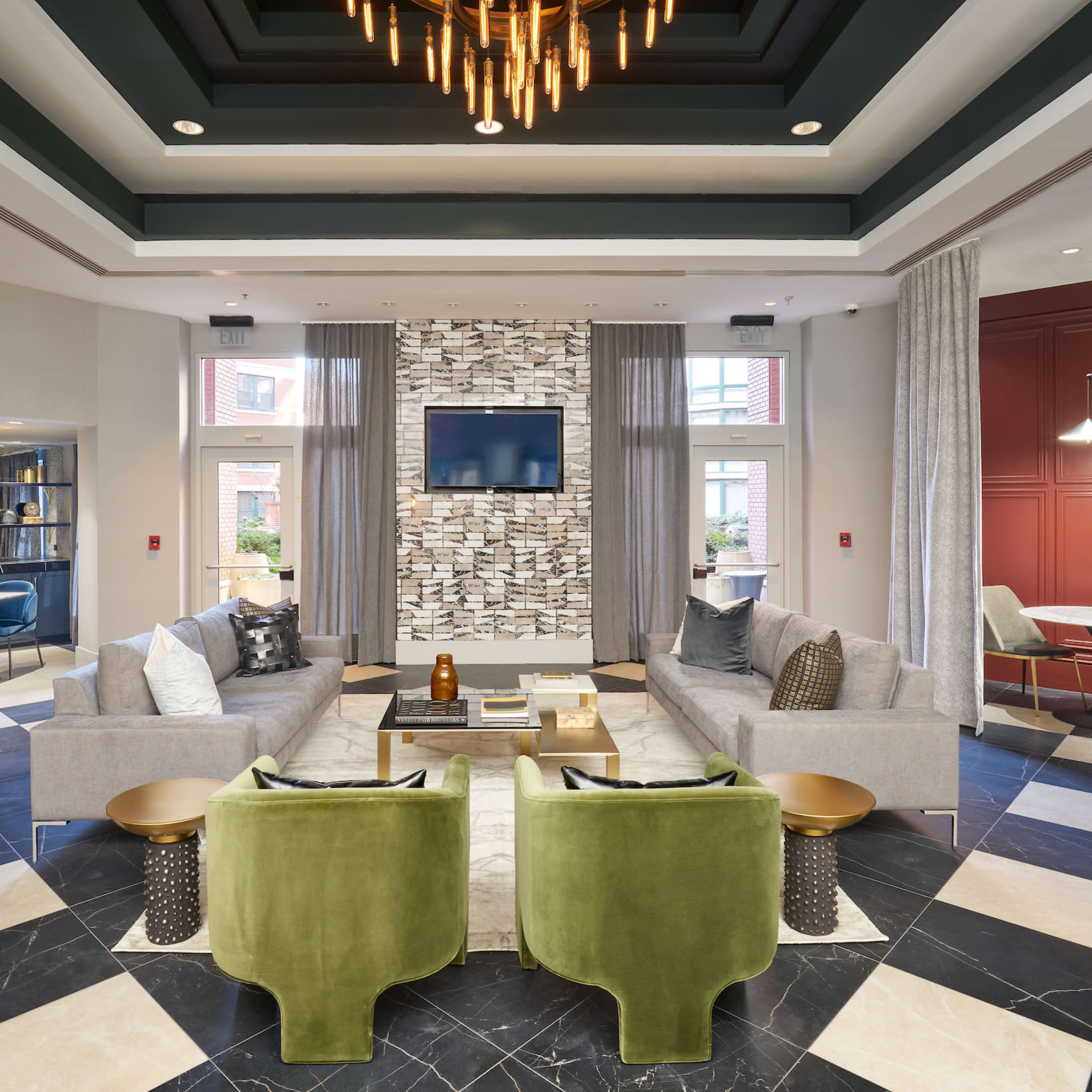 A modern lounge area featuring two green armchairs, a gray sectional sofa, a marble floor with geometric patterns, and a wall-mounted television surrounded by decorative stone.