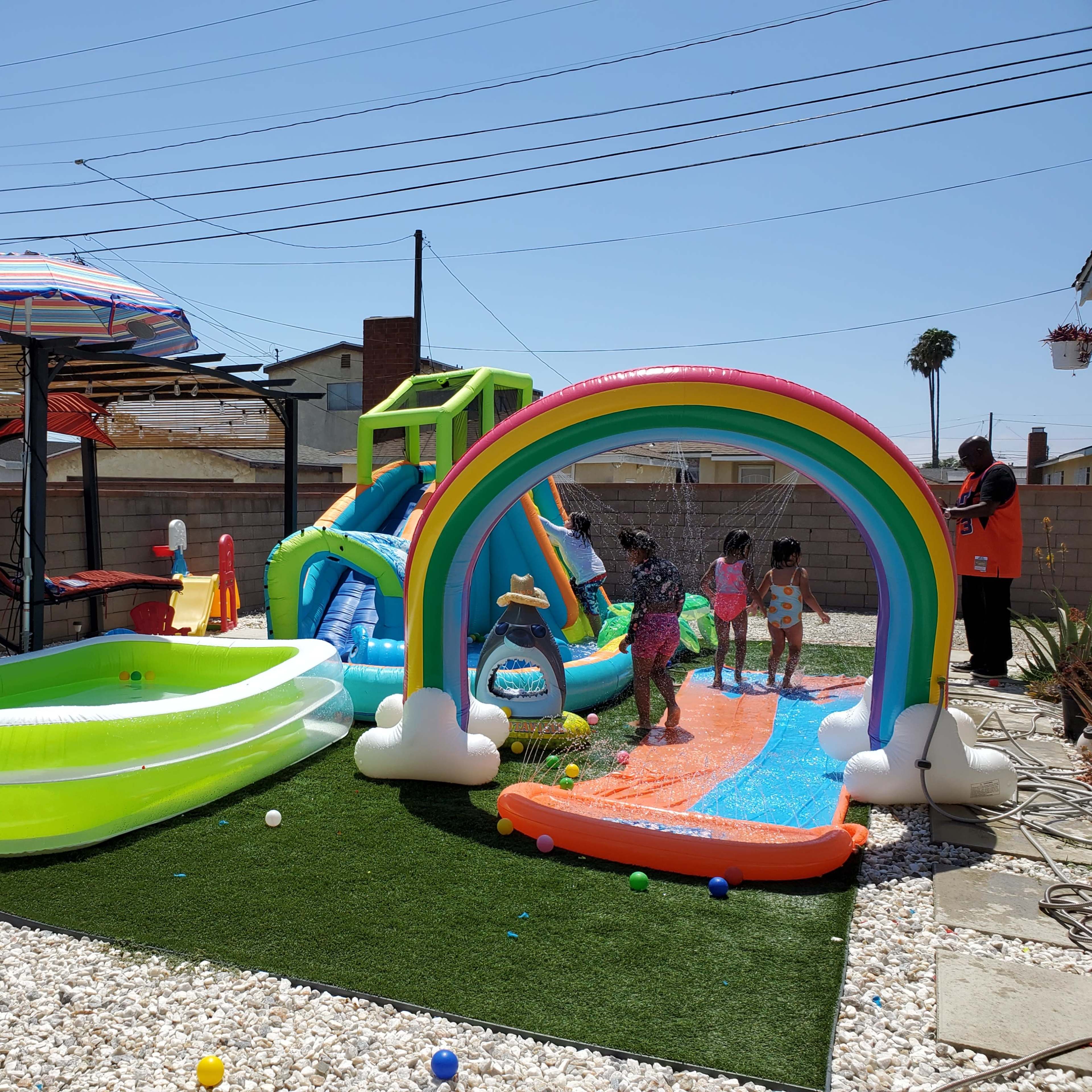 A backyard with a colorful inflatable water slide, a small pool, and children playing on a water-filled slide under a clear blue sky.