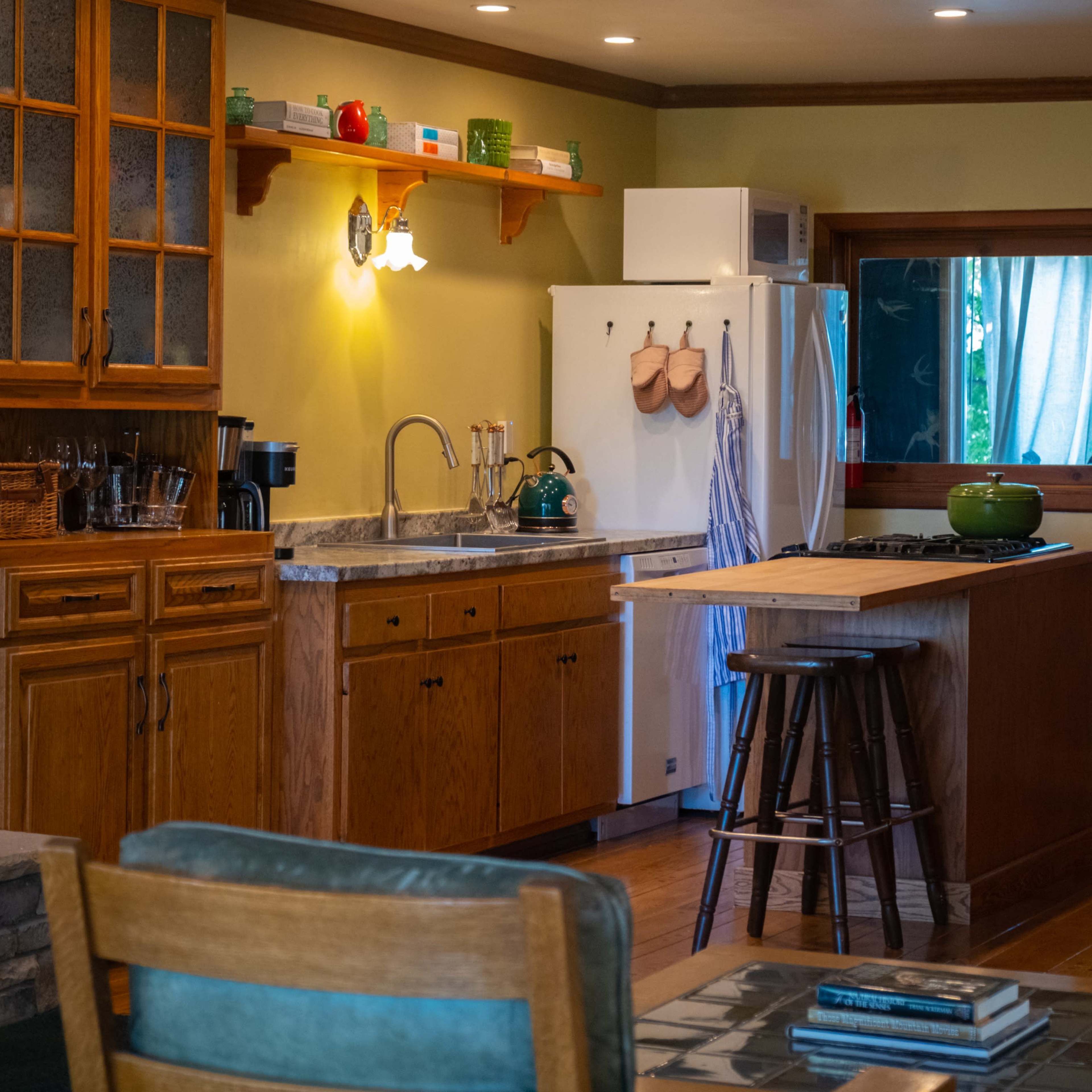 A well-lit kitchen features wooden cabinets, a central island with stools, and a white refrigerator near a window.
