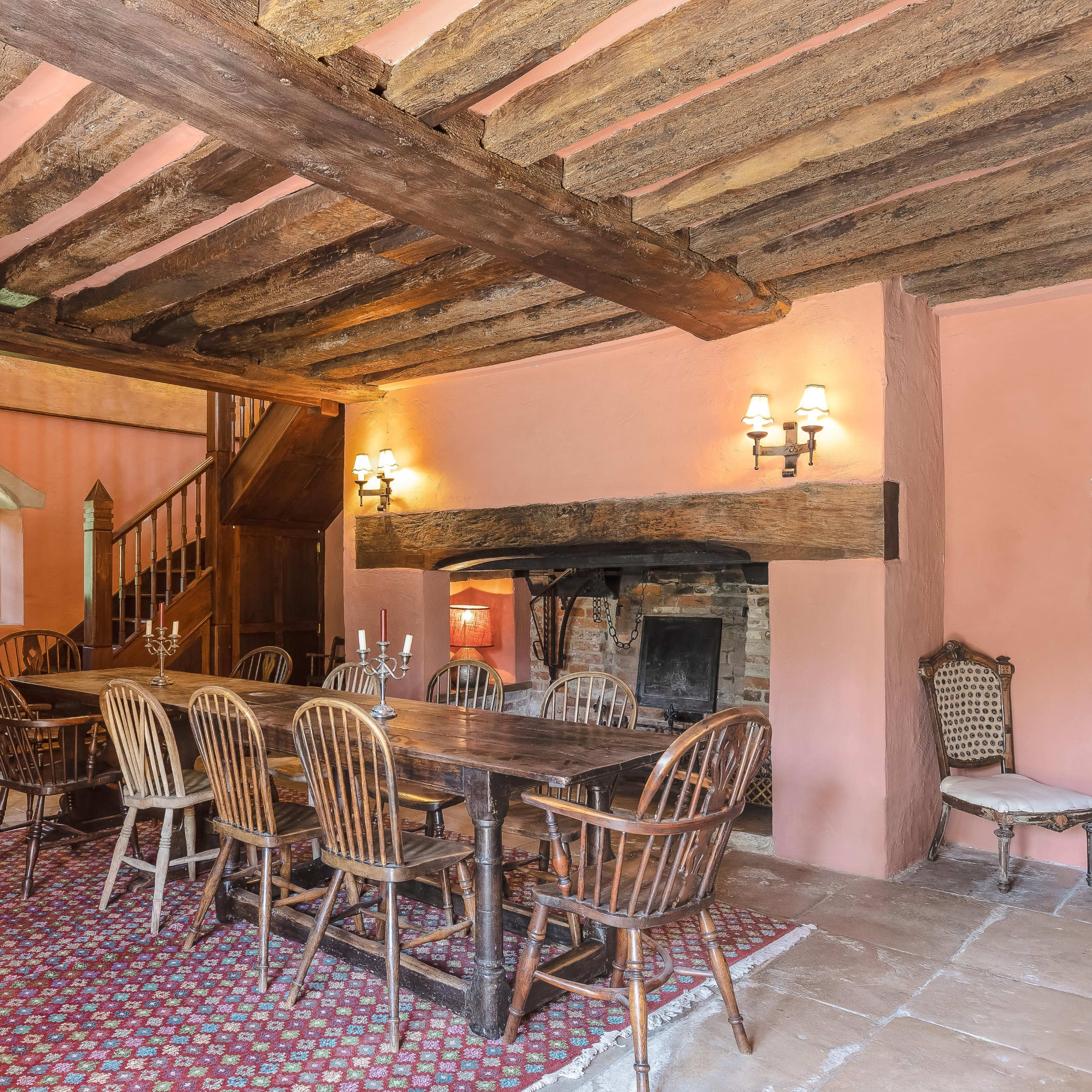 A rustic dining room featuring a long wooden table surrounded by chairs, with exposed beams on the ceiling and a stone fireplace.