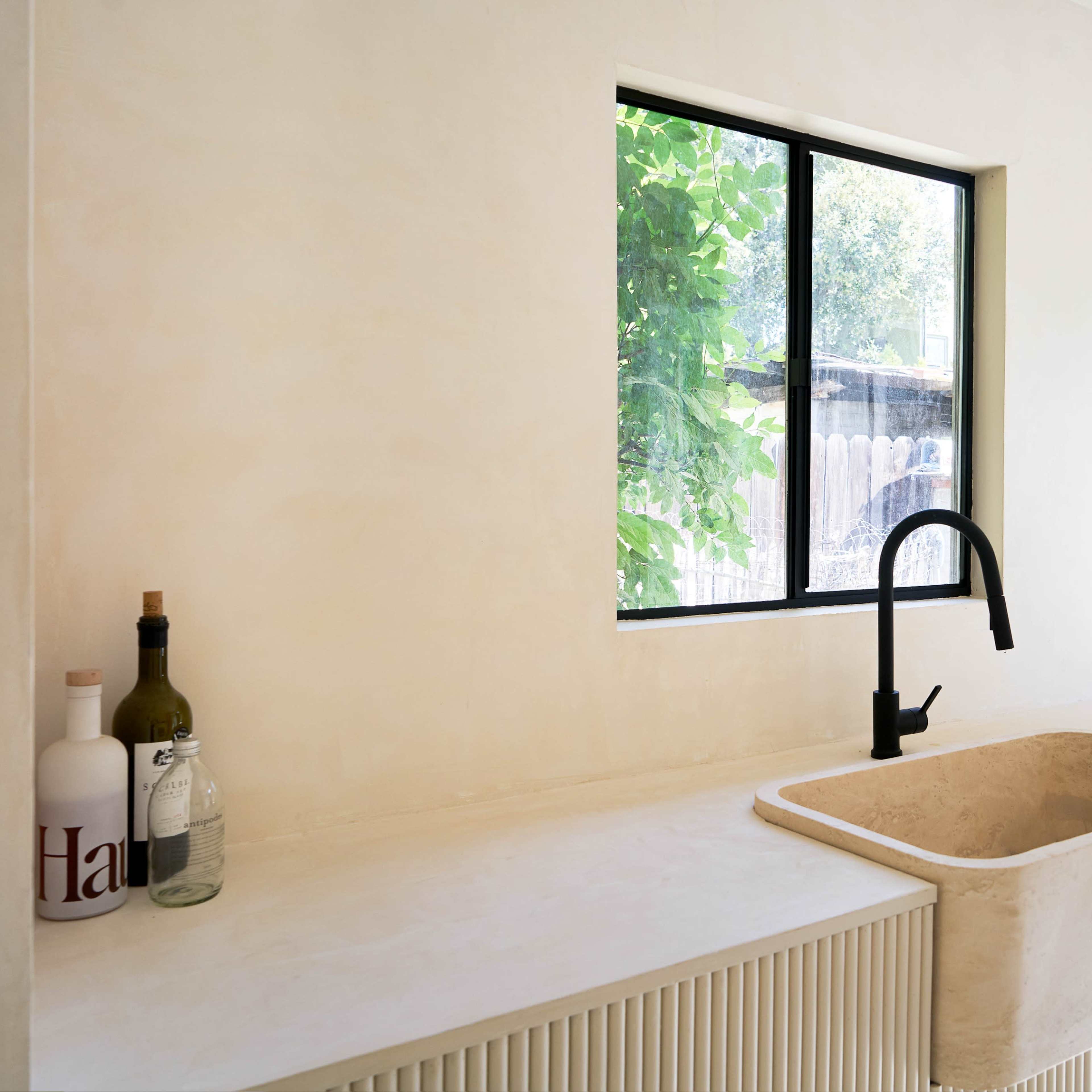 A minimalist kitchen with a large sink and a sleek black faucet, illuminated by natural light through a window.