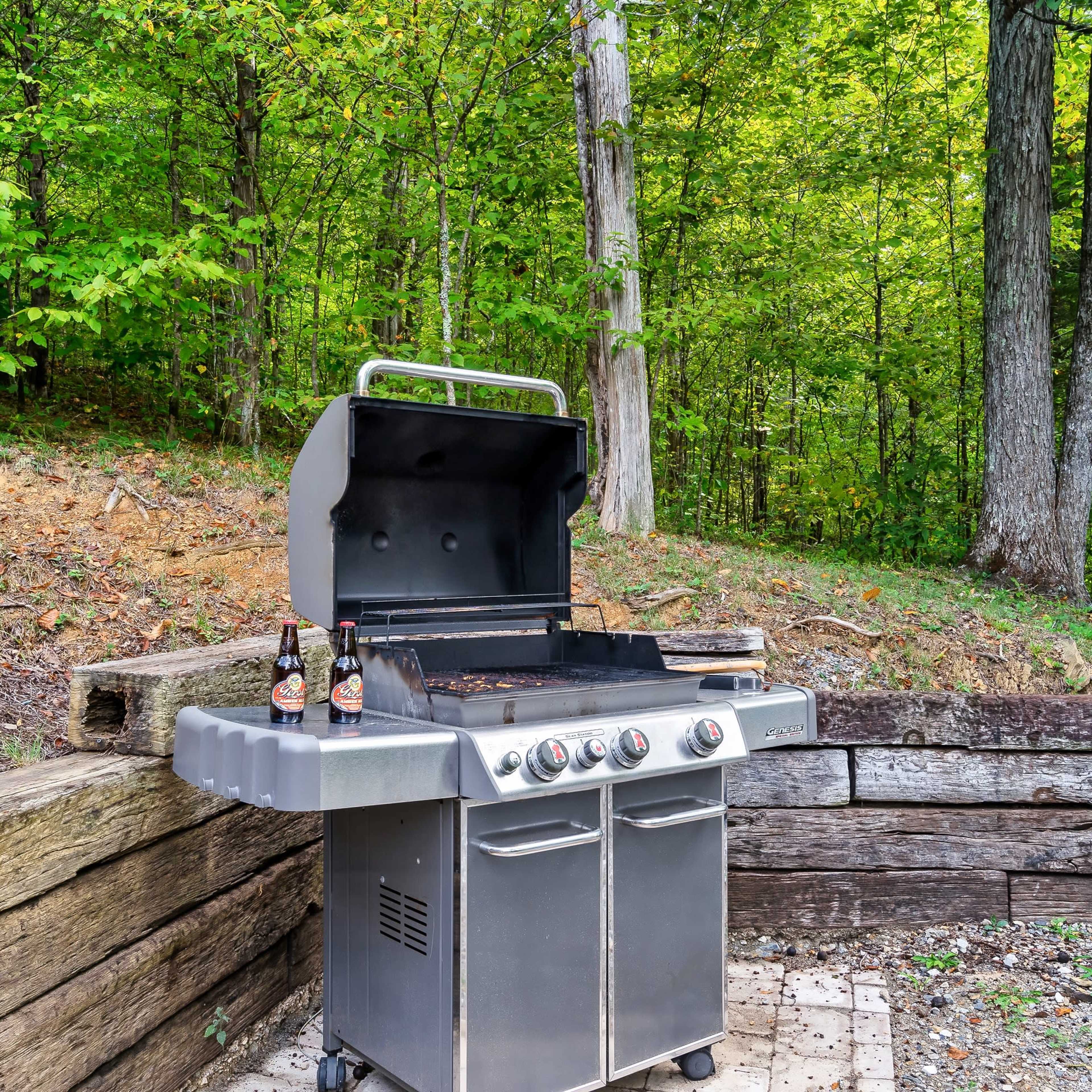 A stainless steel grill with the lid open sits on a stone patio surrounded by trees and gravel.