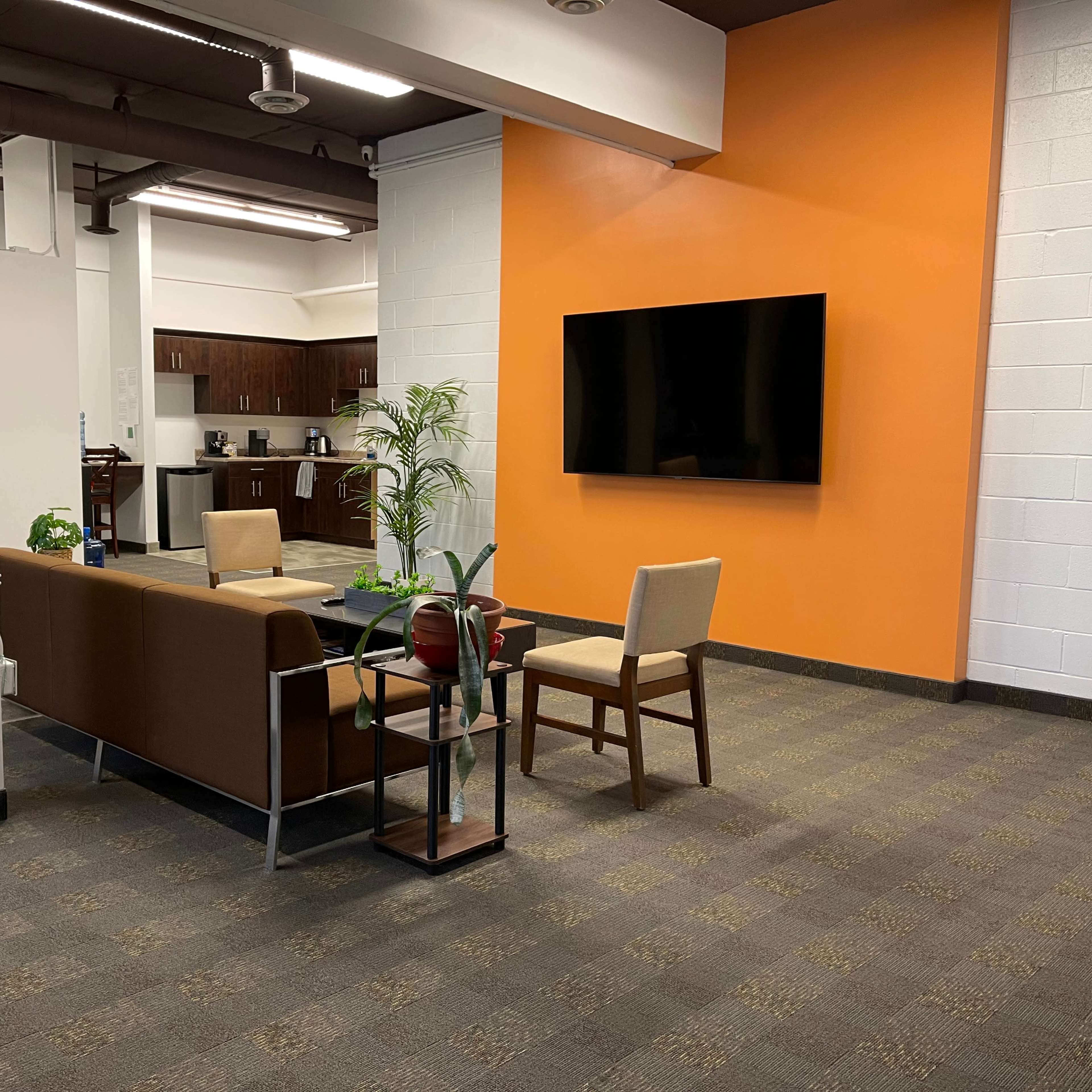 A modern lounge area with a brown sofa, a chair, a small table, and a wall-mounted television, set against an orange accent wall.