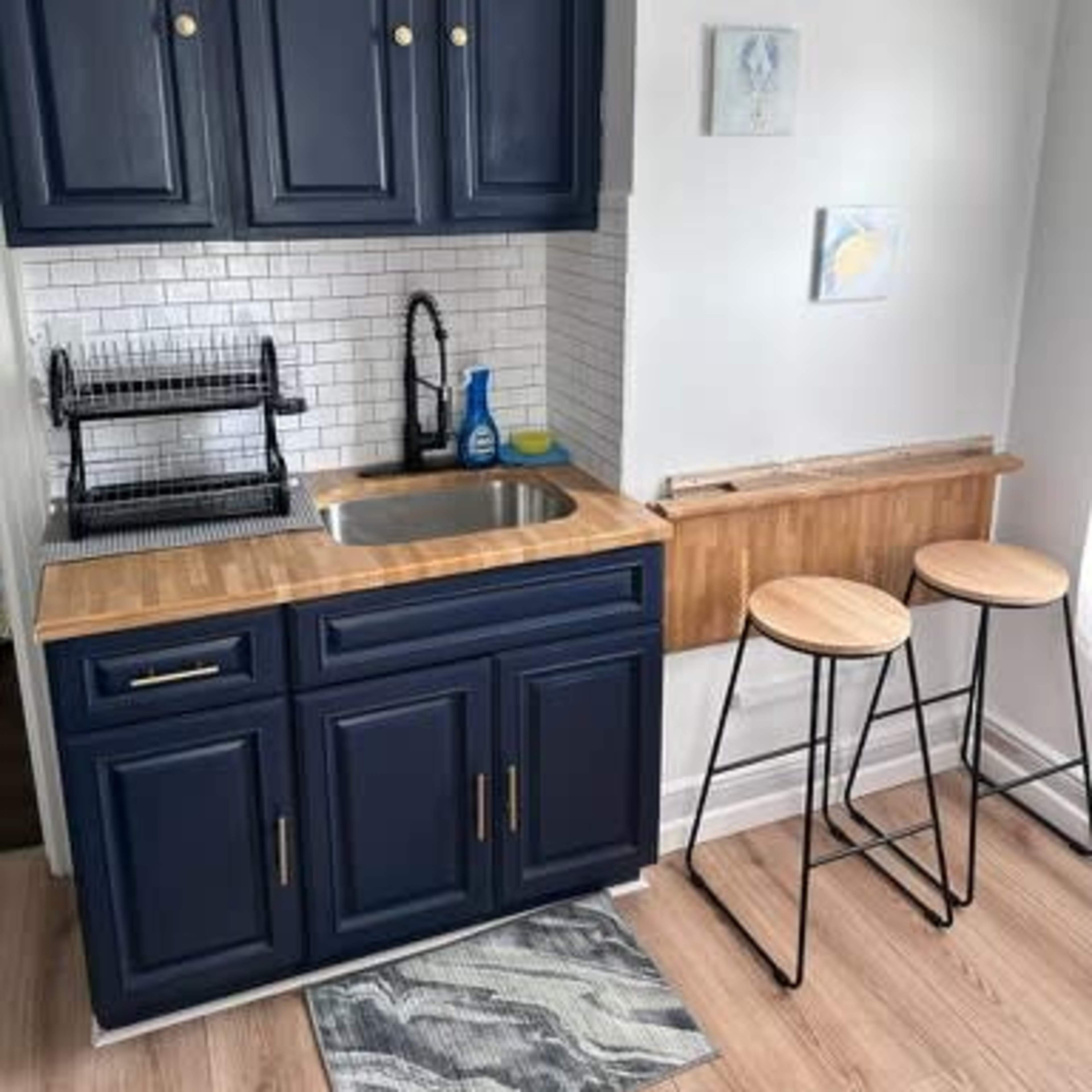 A compact kitchen area featuring navy blue cabinets, a wooden countertop with a sink, and two bar stools next to a small folding table.