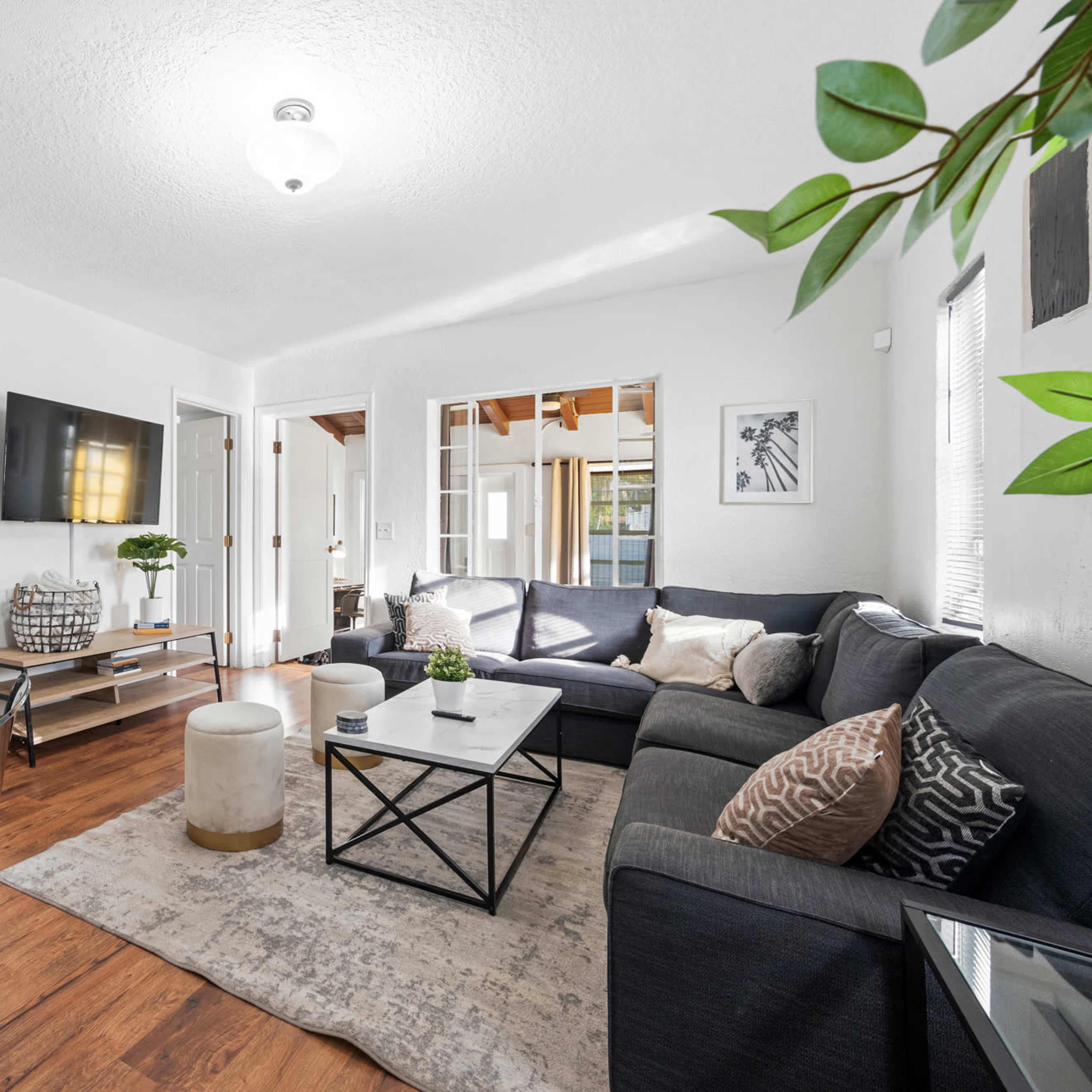 A modern living room features a dark sectional sofa, a coffee table, and a dining area with a glass table, accented by natural light and greenery.