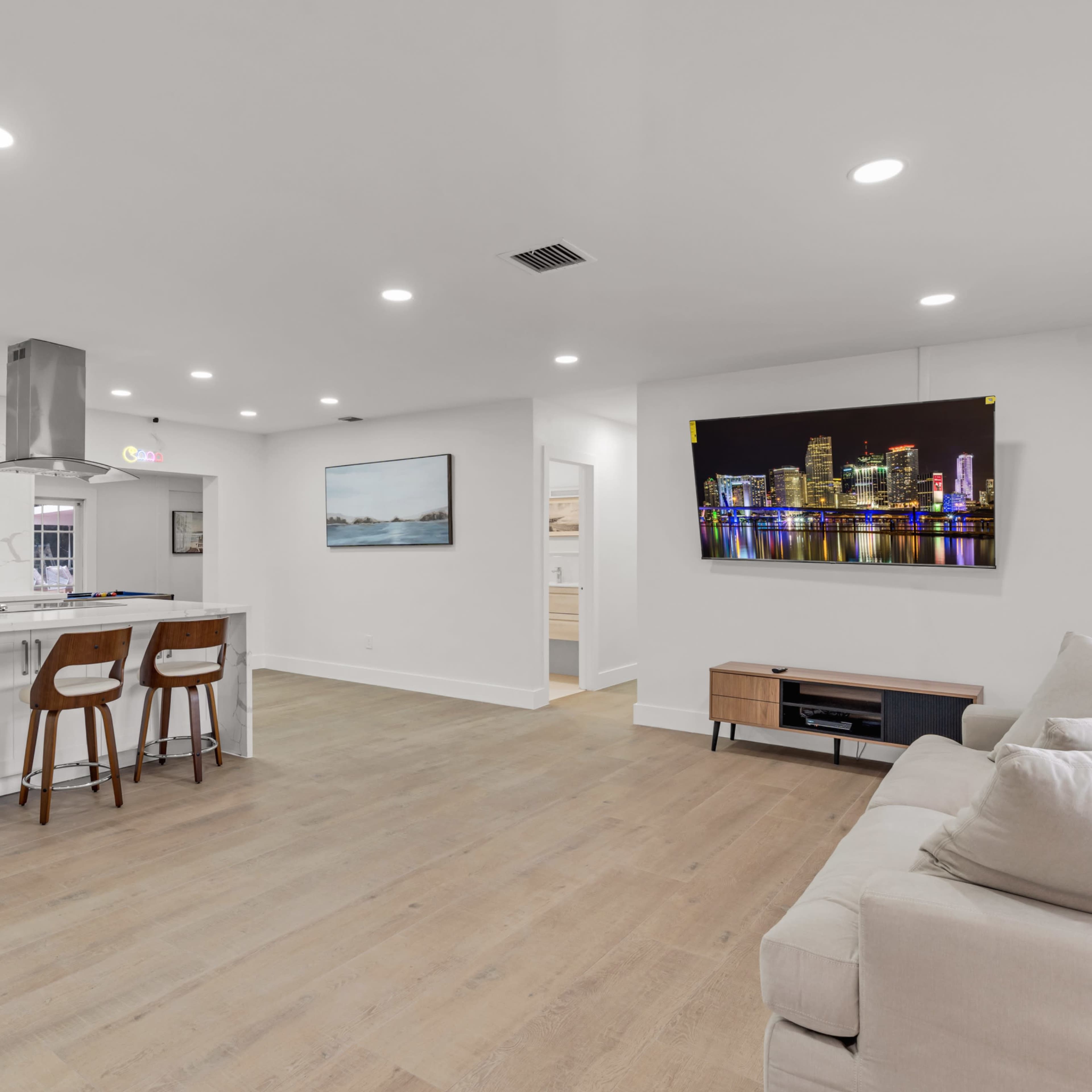A modern open-concept living space featuring a kitchen with a large island, two bar stools, and a living area with a couch and a wall-mounted television displaying a city skyline.