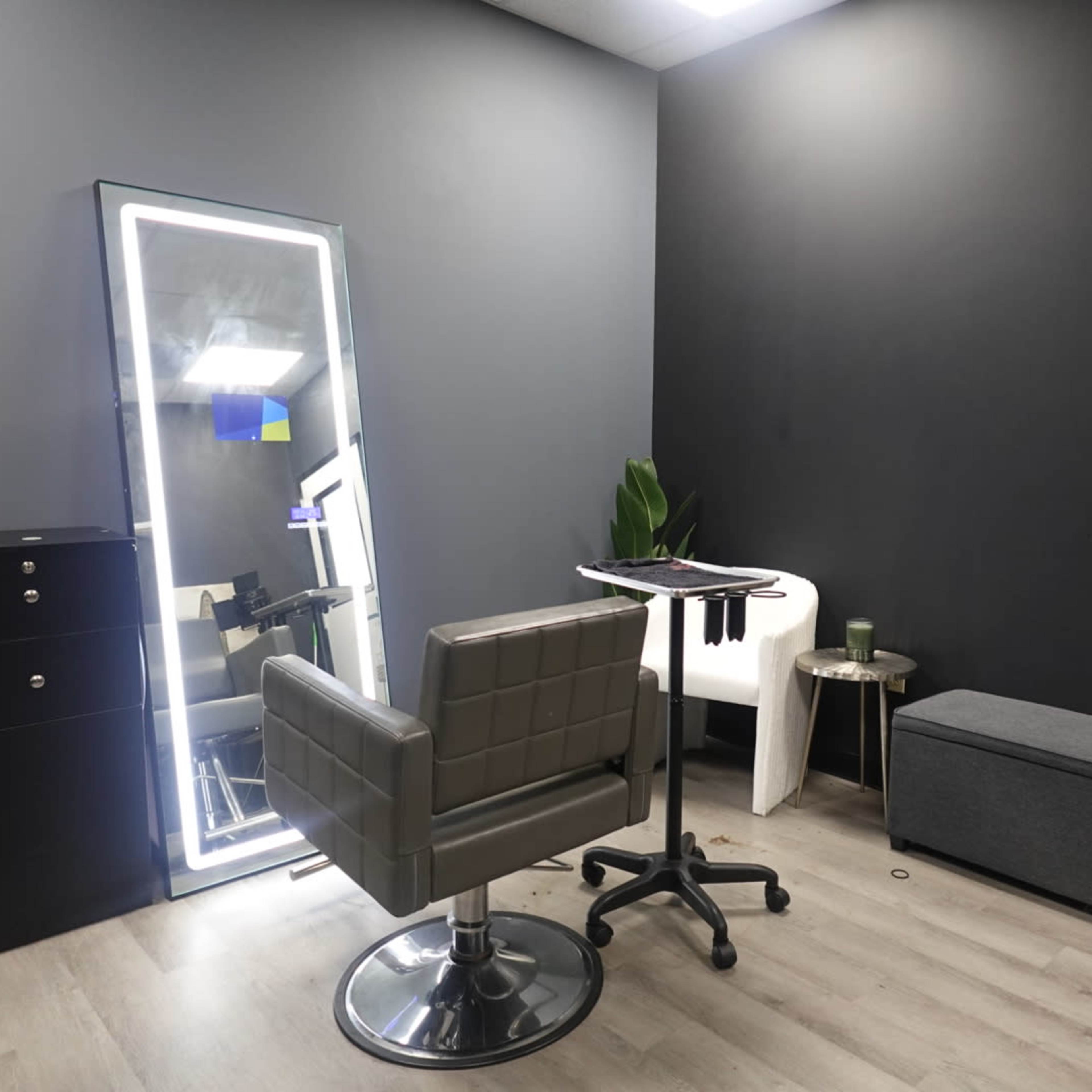 A modern hair salon interior featuring a stylist chair, a tall mirror, a small table with tools, and storage units against dark walls.
