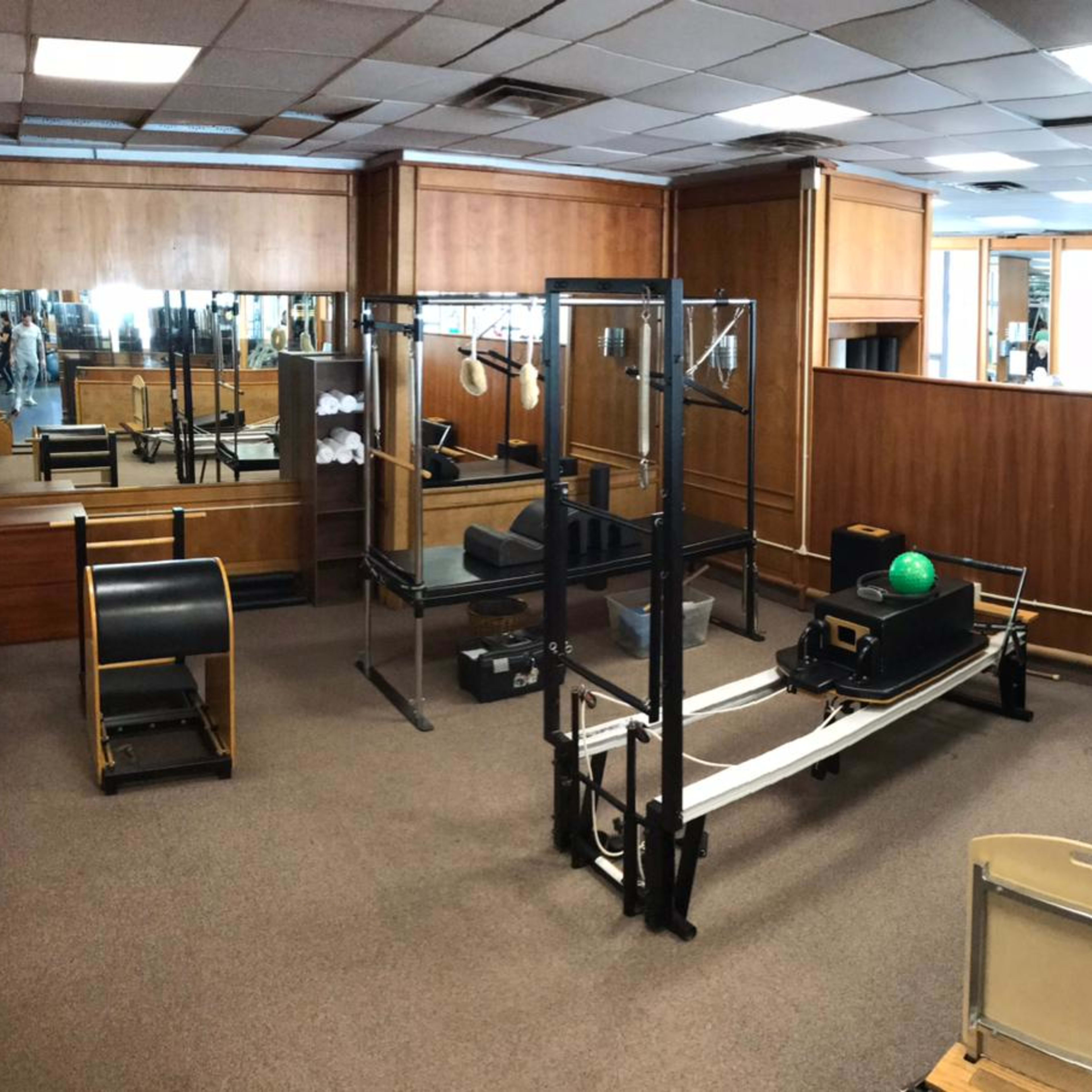 A well-lit Pilates studio equipped with various exercise machines, including a reformer and a cadillac, set against wooden paneling and mirrors.