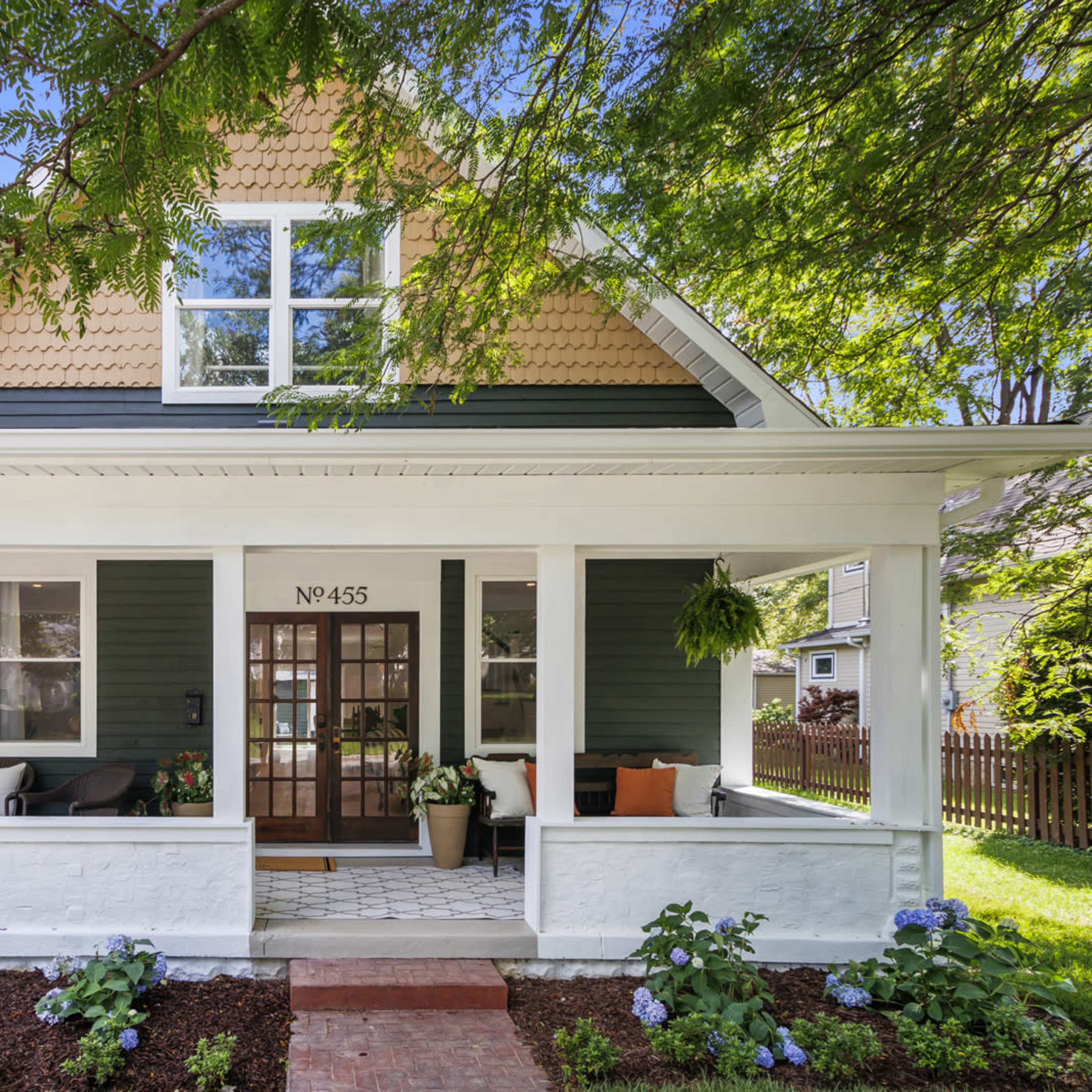 A two-story house with a green exterior, white trim, and a front porch decorated with potted plants and seating.