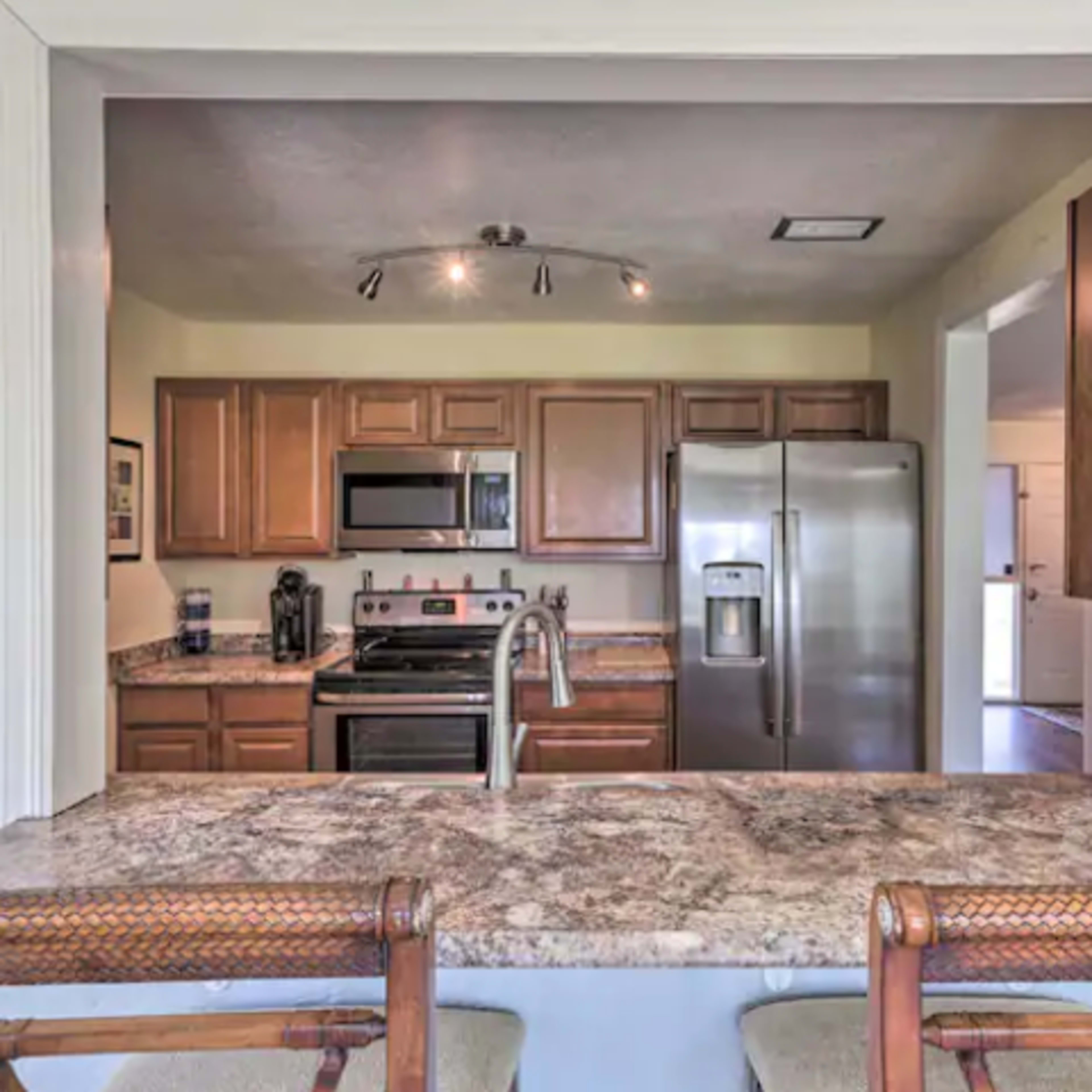 A kitchen viewed from a bar area, featuring wooden cabinets, stainless steel appliances, and a granite countertop.