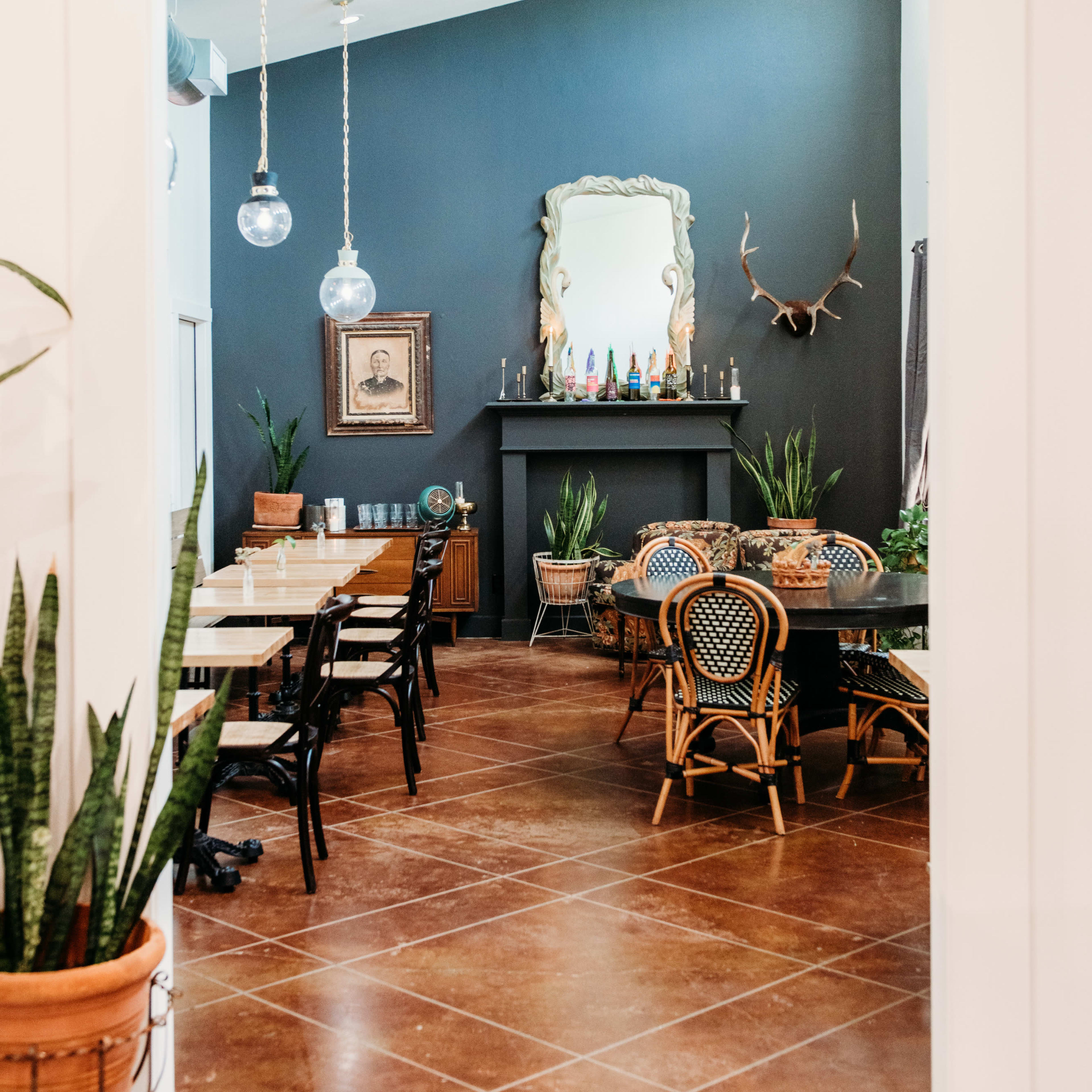 A dining area with wooden tables and chairs, decorative plants, and a dark wall featuring a mirror and antlers.