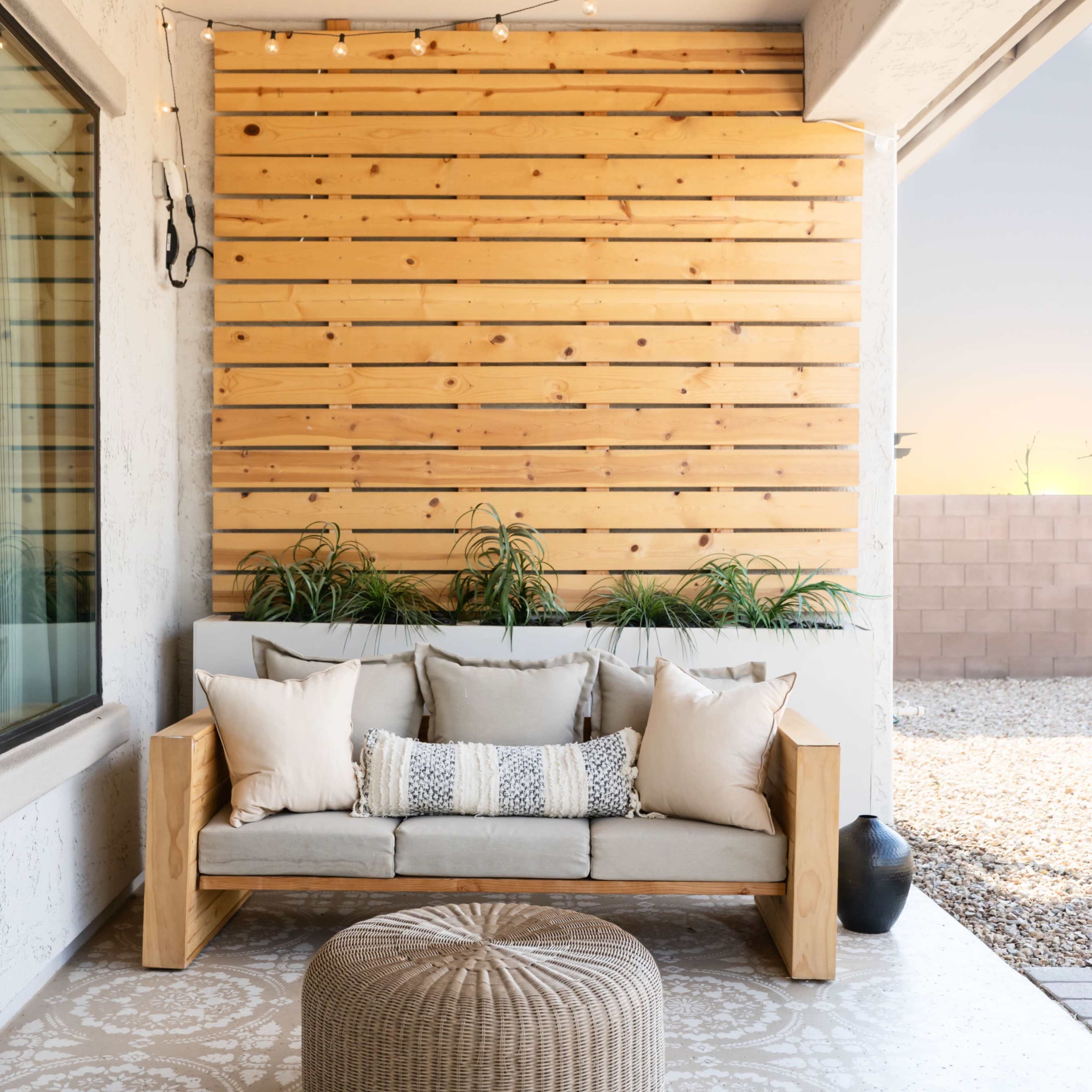 A wooden sofa with cushions is positioned on a patio beneath string lights, next to a round pouf and a decorative planter against a slatted wood wall.