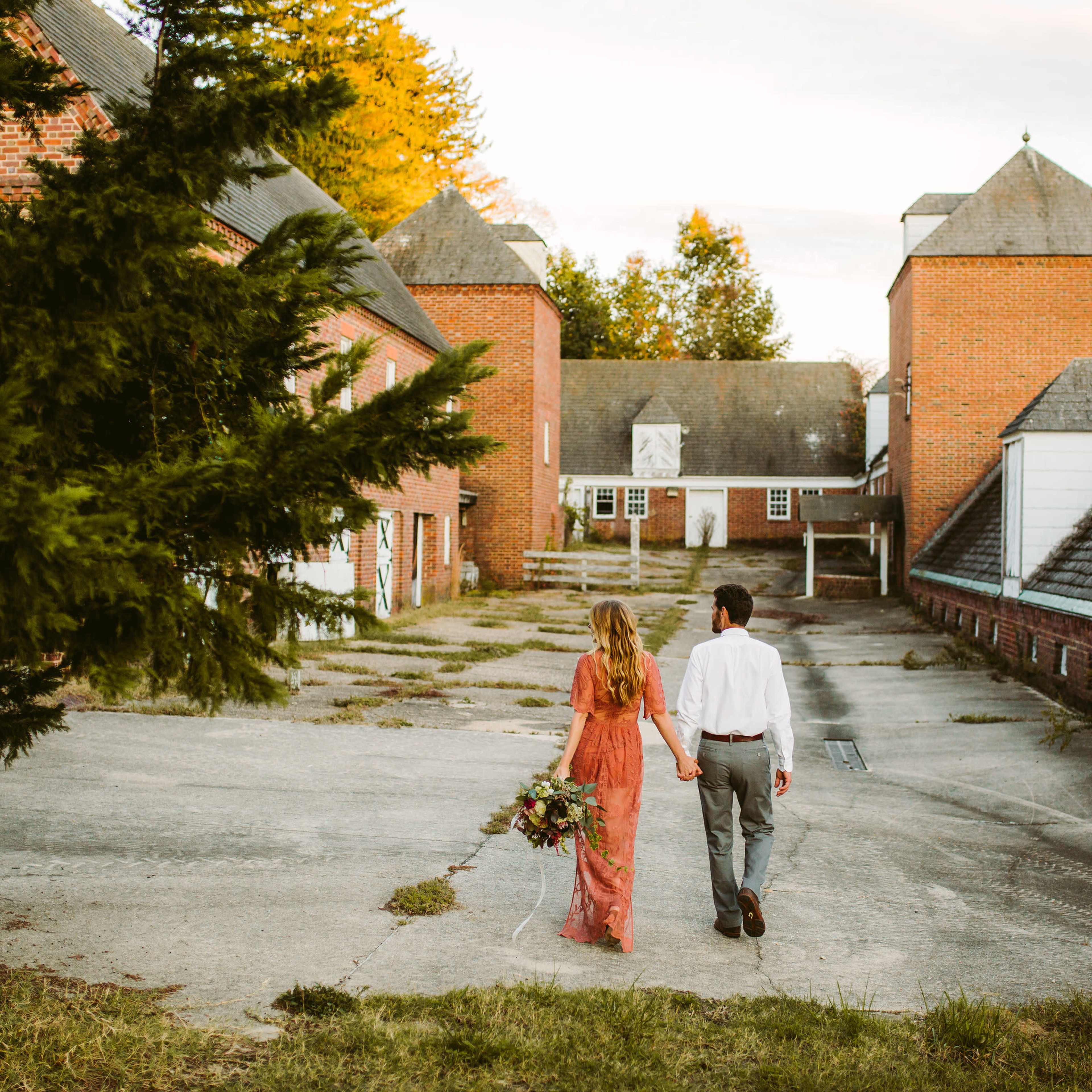 A couple walks hand in hand through a landscaped area with brick buildings in the background.