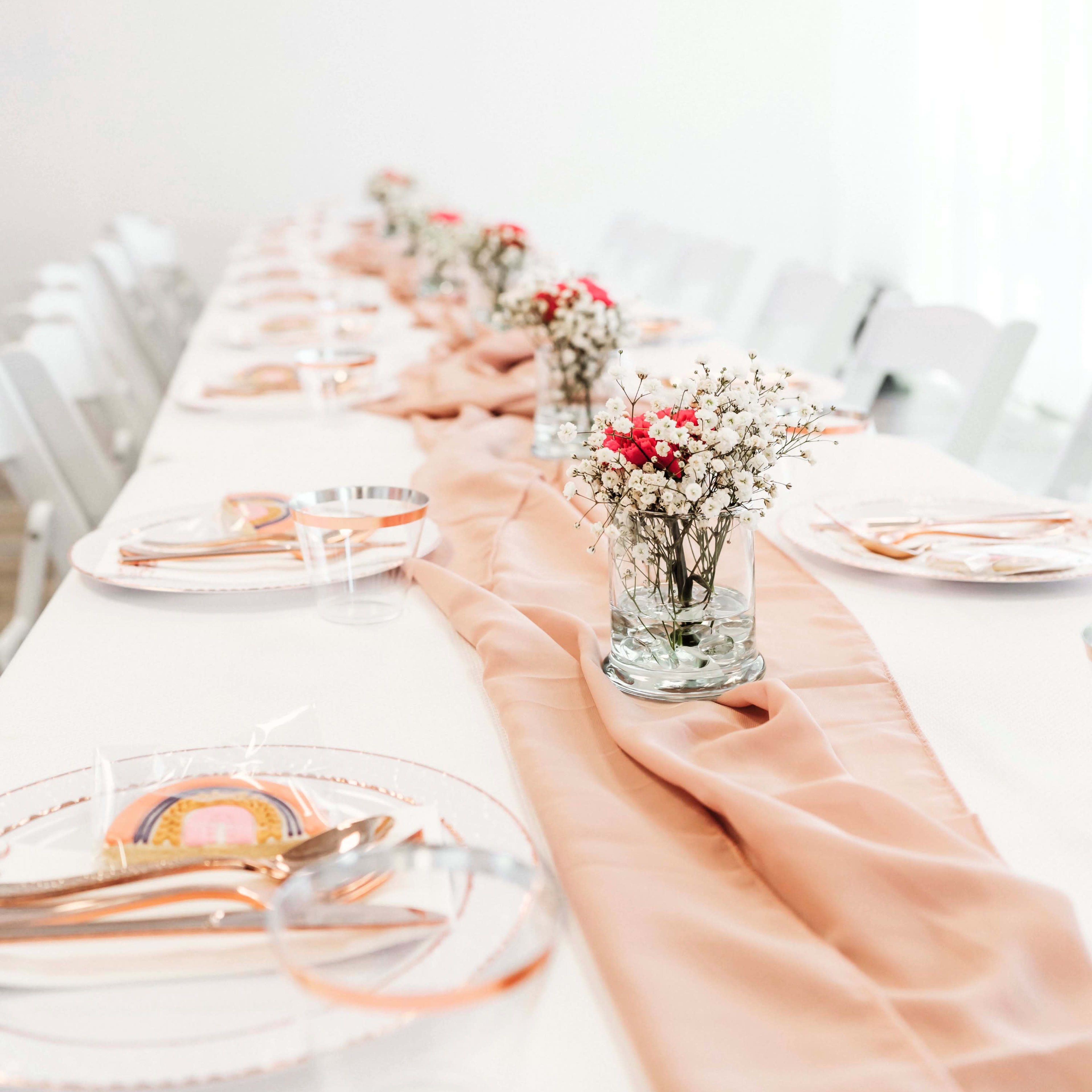 A long dining table is set with clear plates, gold utensils, and a light peach table runner, adorned with small floral centerpieces in glass vases.