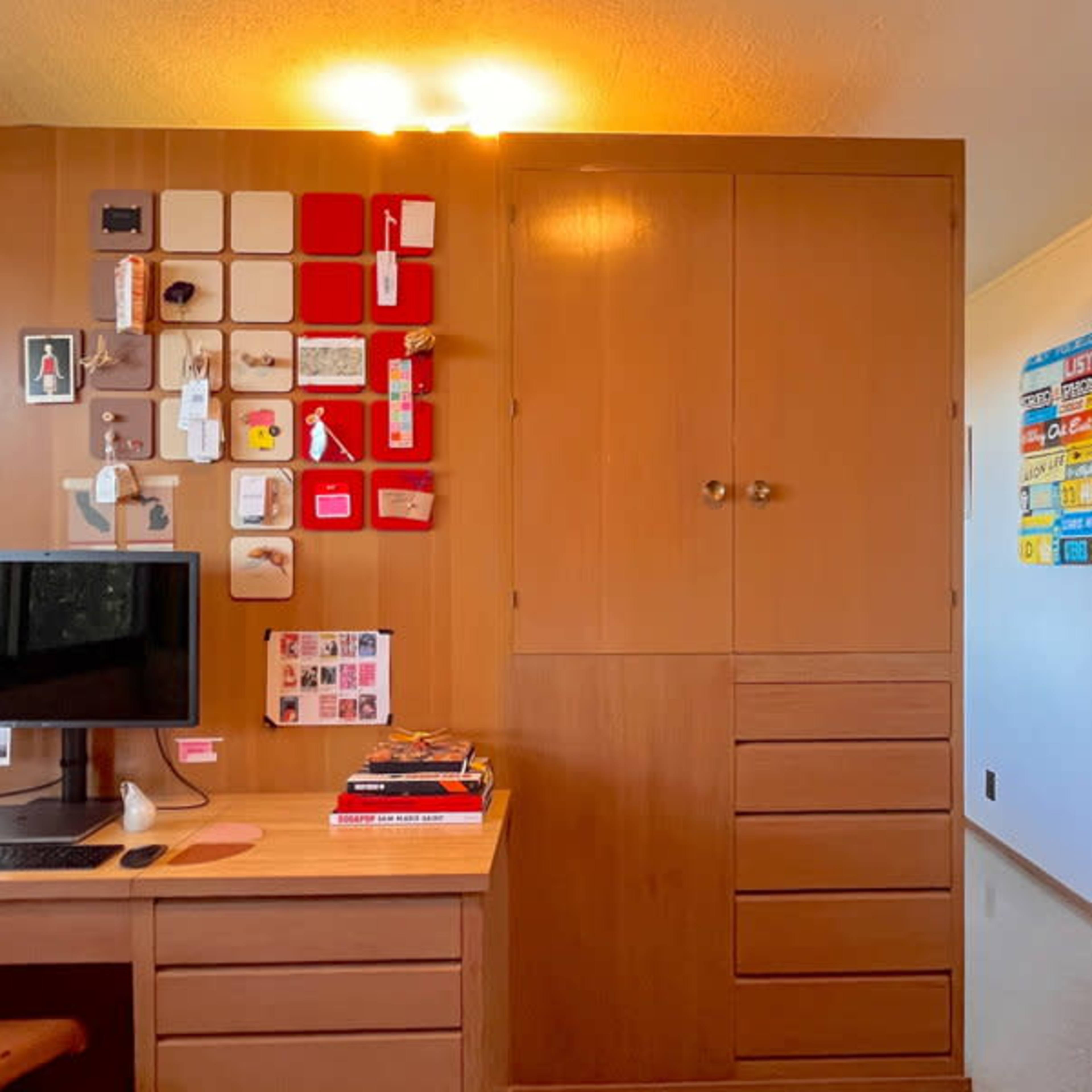 A wooden desk with a computer and various items on the surface, adjacent to a wall featuring a grid of colorful squares and storage cabinets.