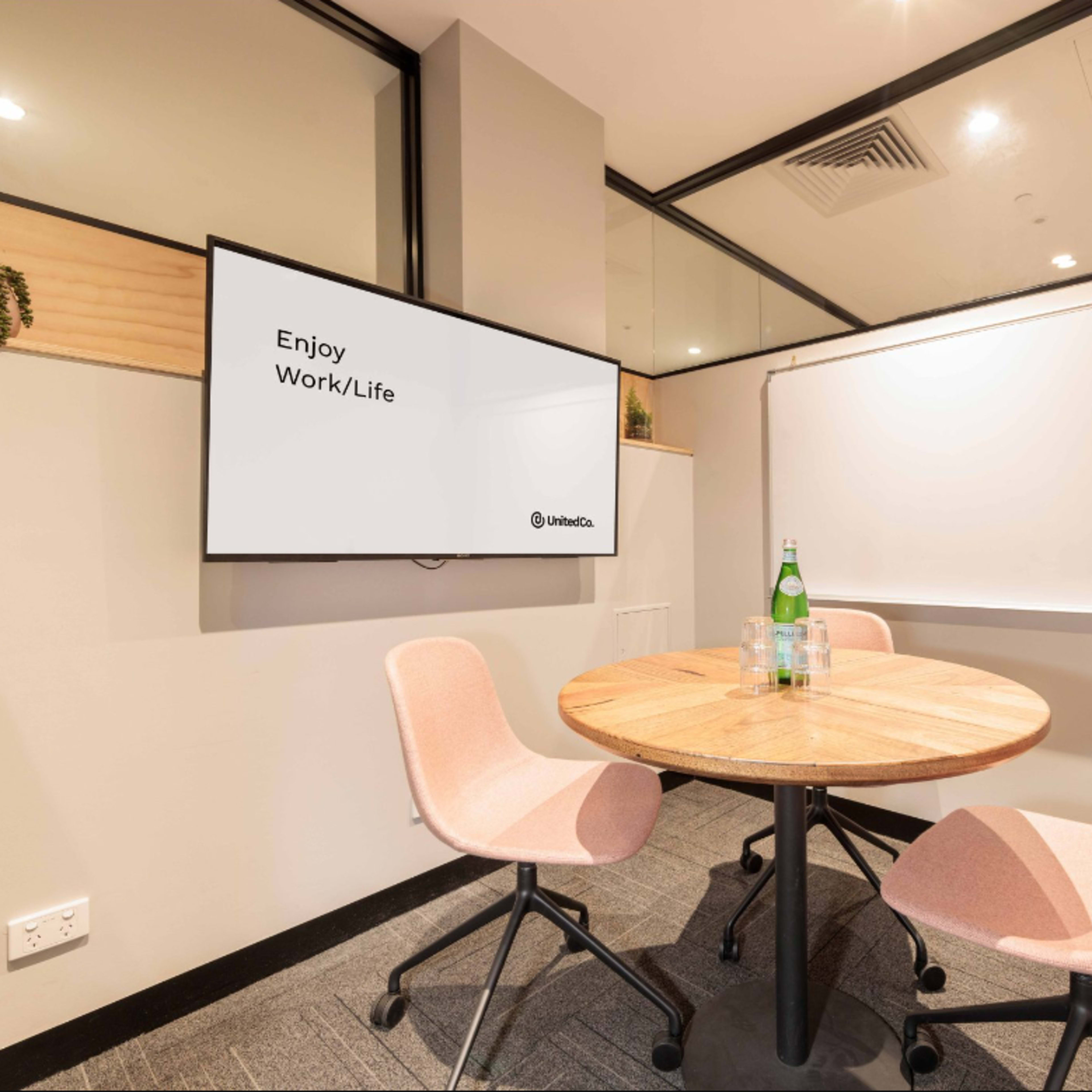A small meeting room features a round wooden table with two pink chairs, a television displaying "Enjoy Work/Life," and a whiteboard.