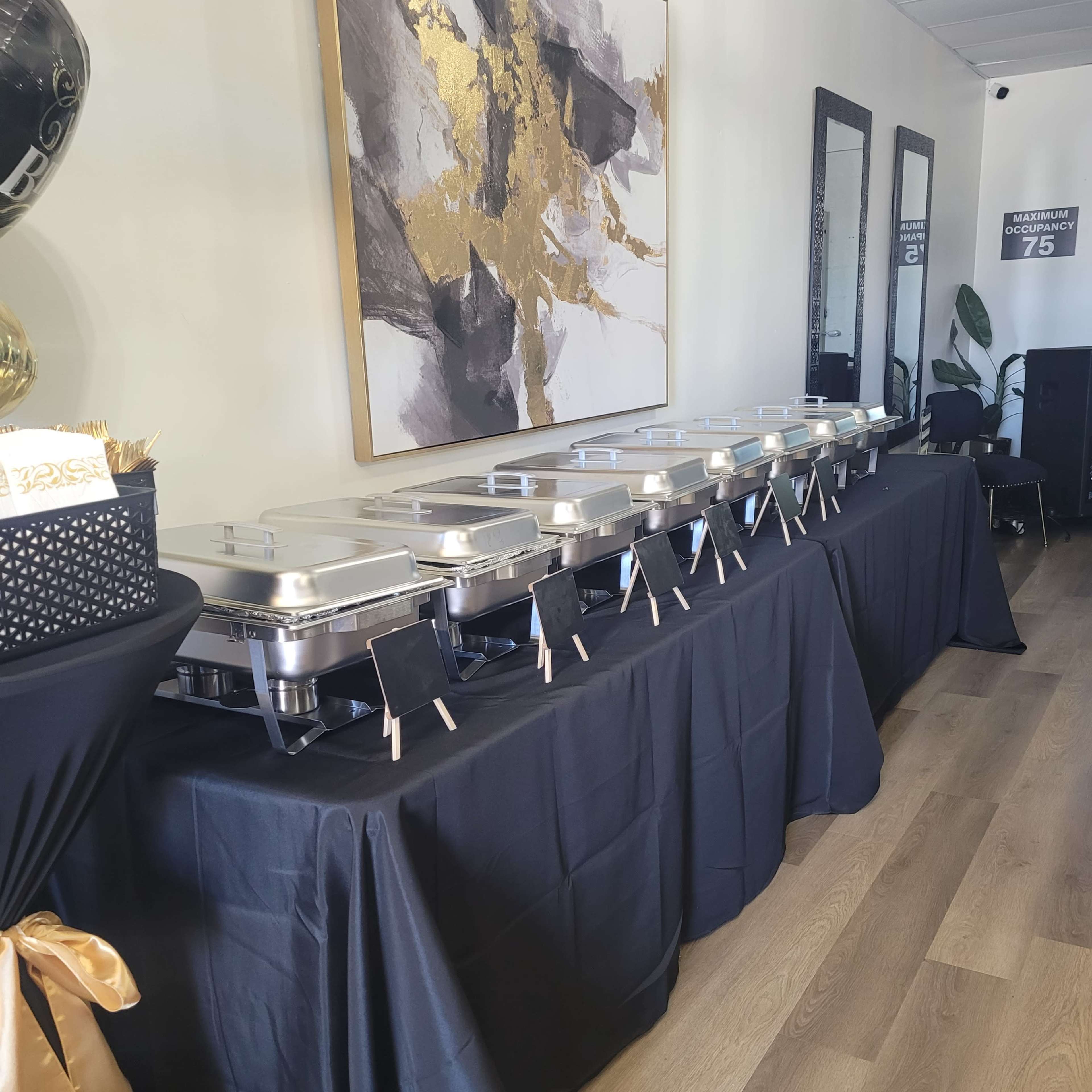 A long table covered with a black tablecloth is set with multiple chafing dishes, and decorative elements are displayed on the surrounding walls.