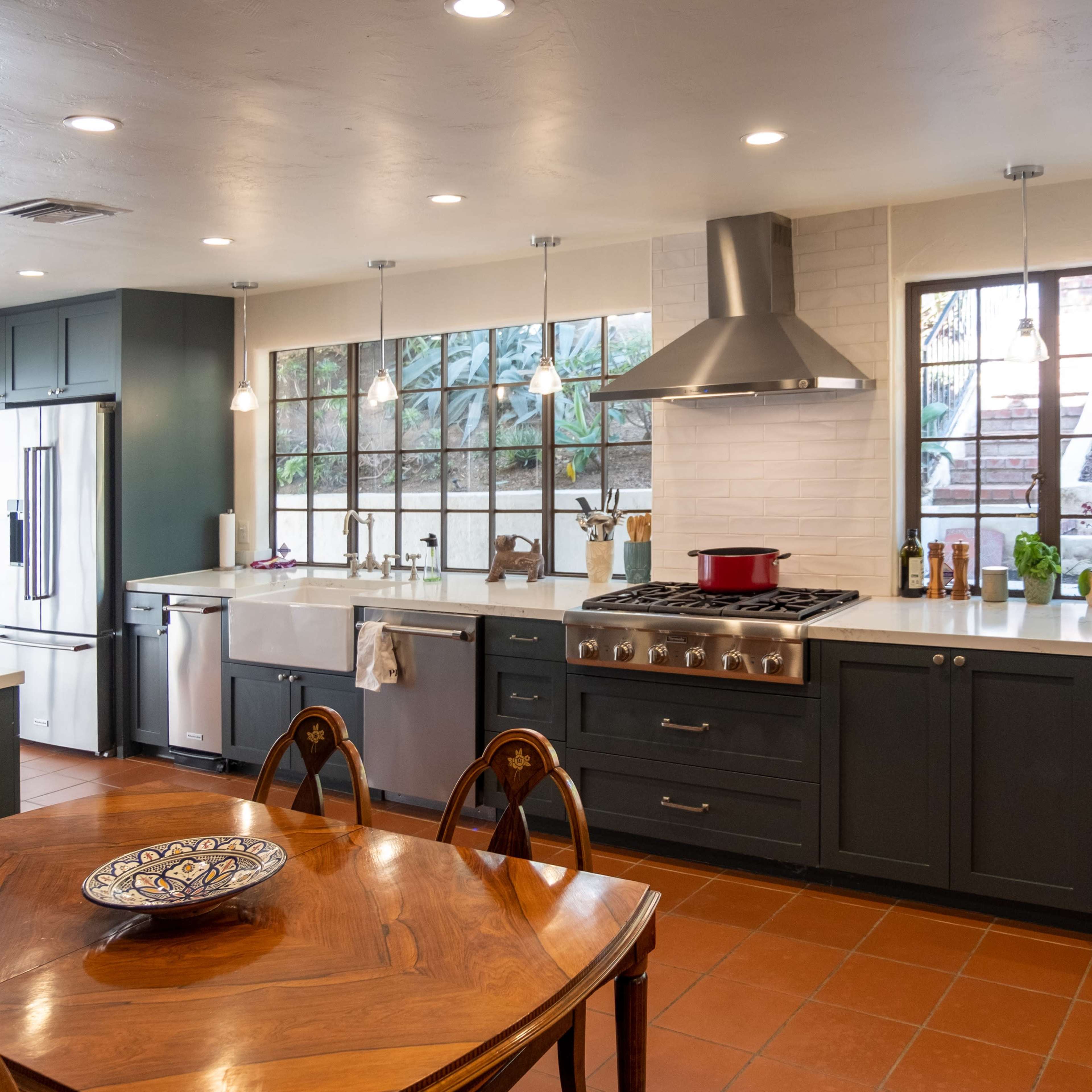 A modern kitchen with dark cabinetry, a large oven, and a dining table set for meals, illuminated by pendant lights and natural light through large windows.