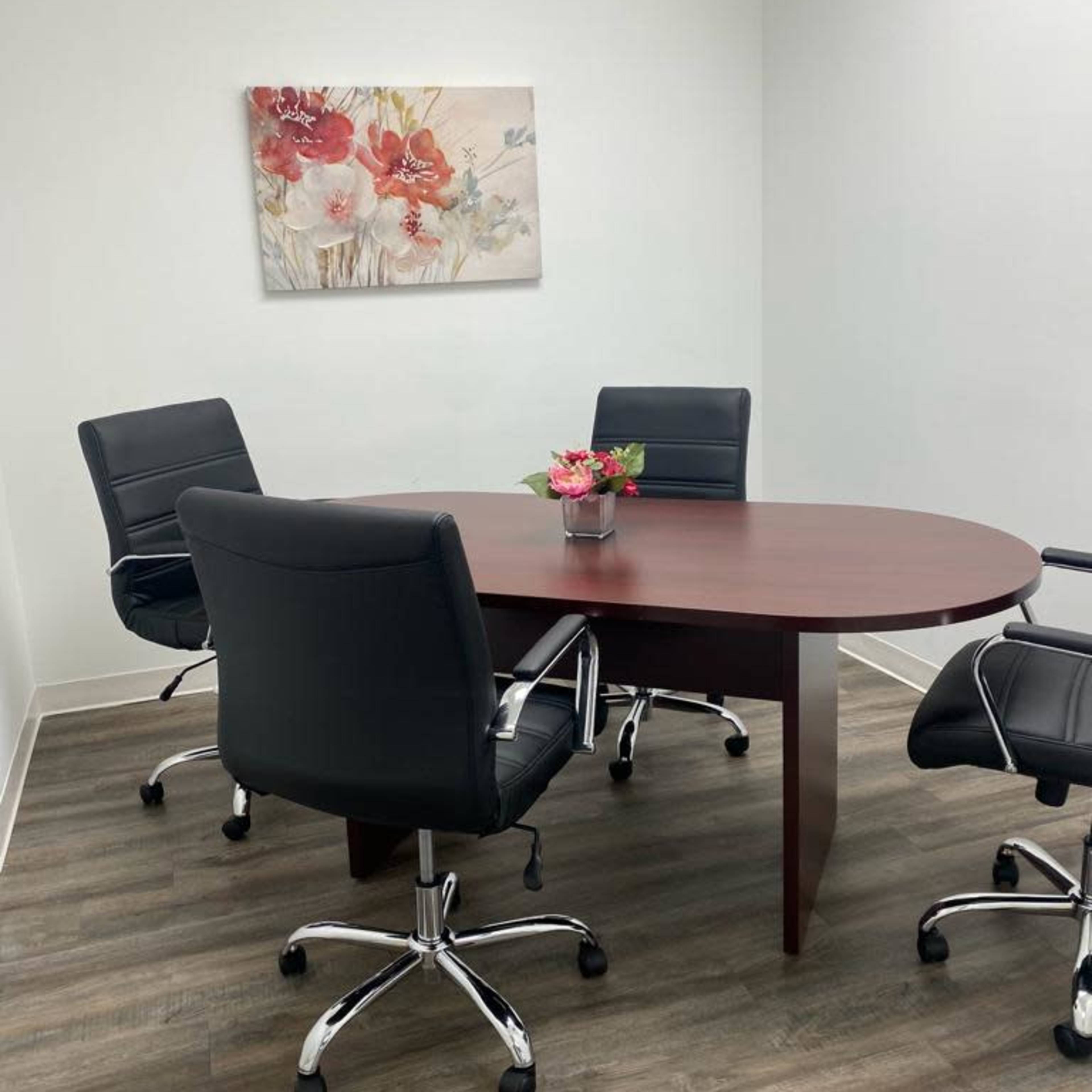 A small conference room with a round table and four black office chairs, decorated with a floral painting on the wall.