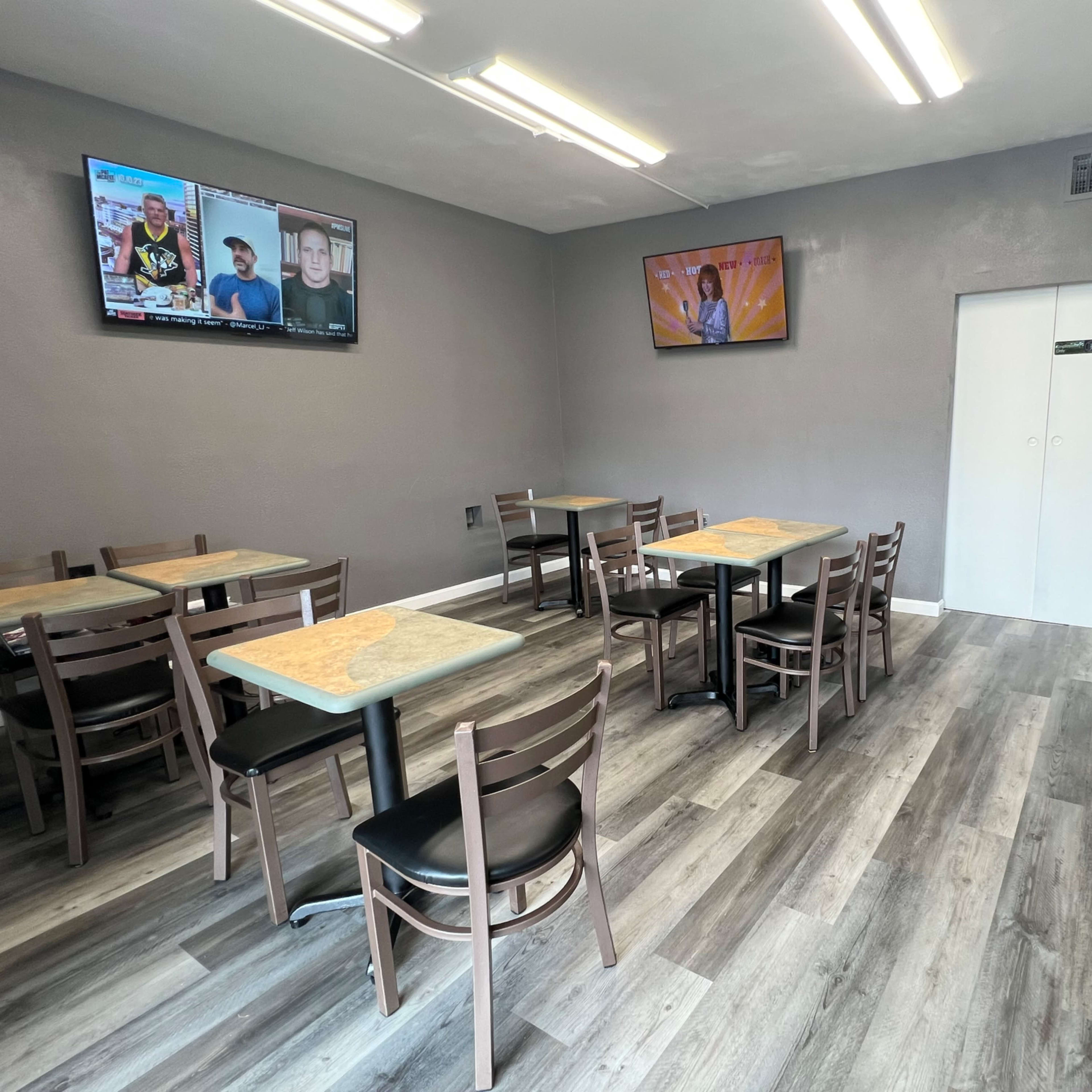 A simple dining area with several tables and chairs, two televisions mounted on the wall, and a microwave next to a storage cart.