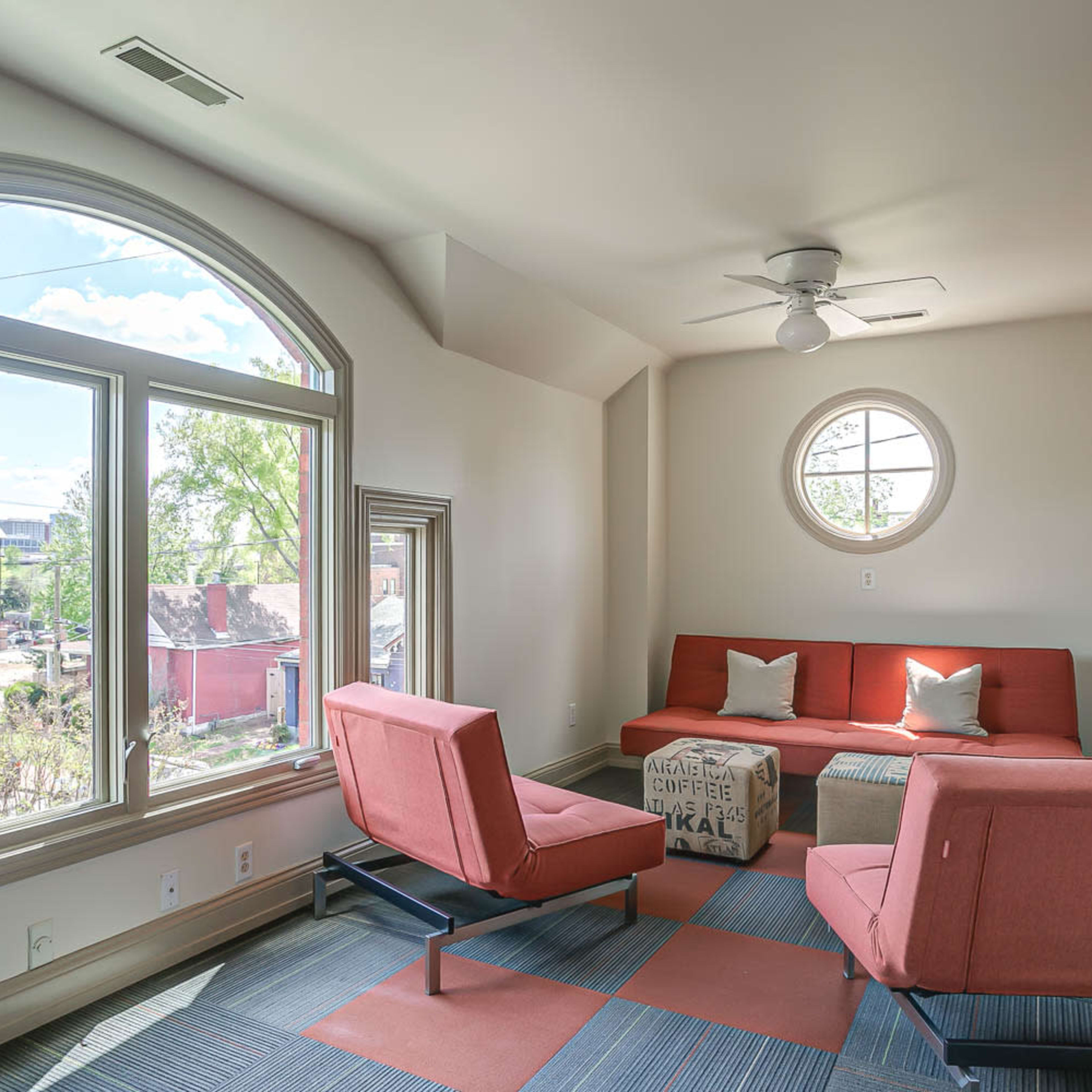 A cozy seating area with two orange chairs and a sofa facing large arched windows that provide a view of the outside.