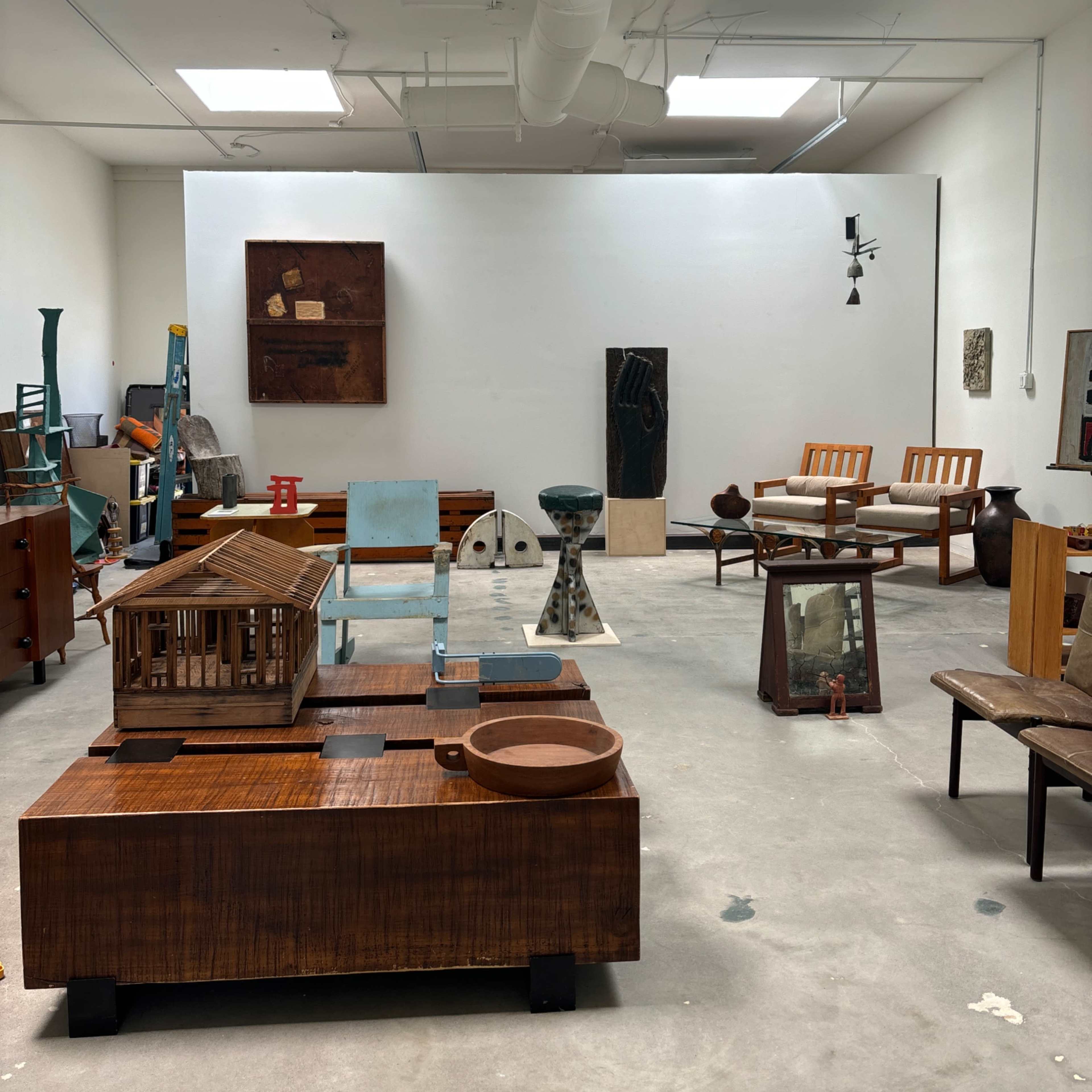 An art gallery or studio featuring a variety of wooden sculptures and furniture pieces arranged on a concrete floor.