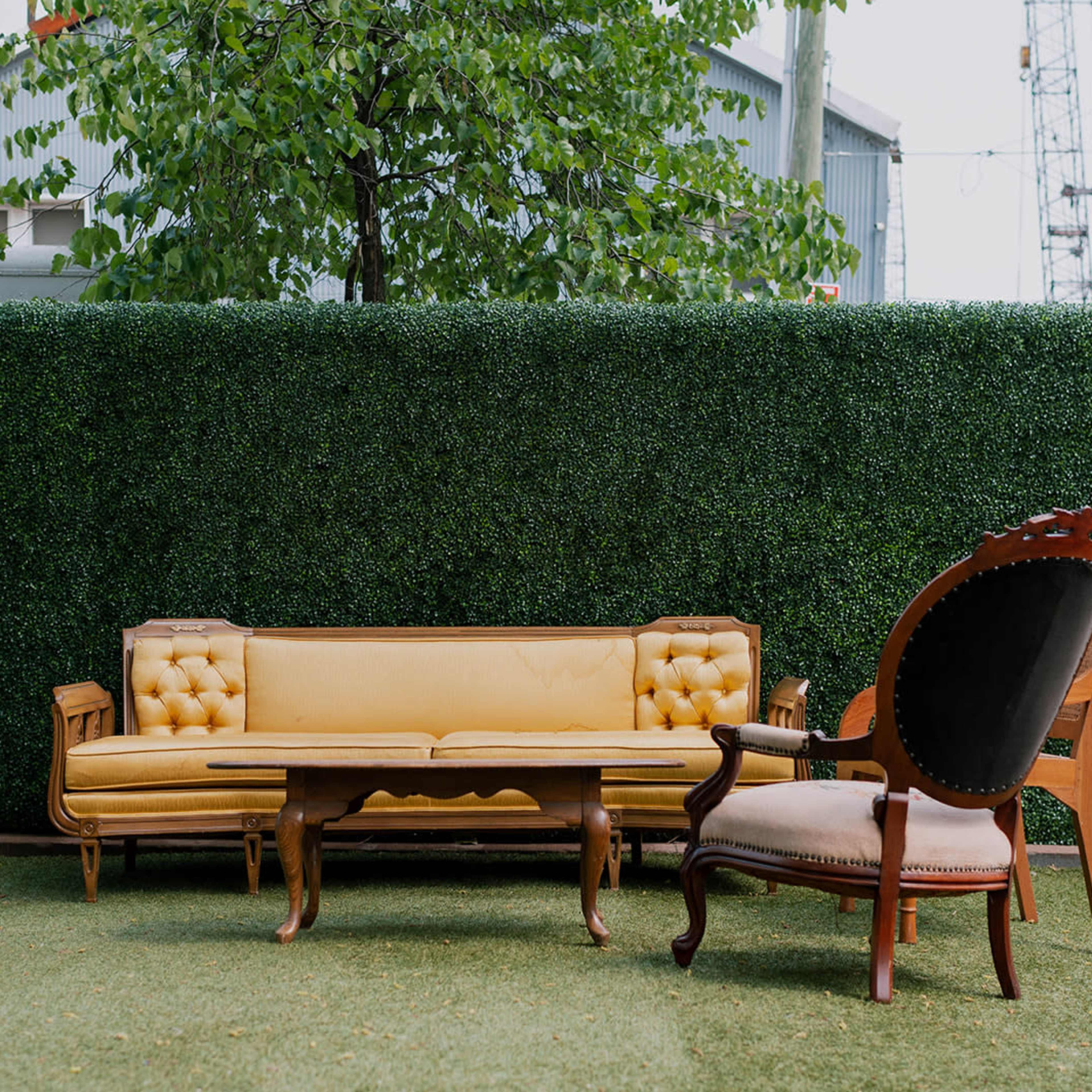 A yellow tufted couch and an ornate chair are positioned on a grassy area surrounded by a green hedge.