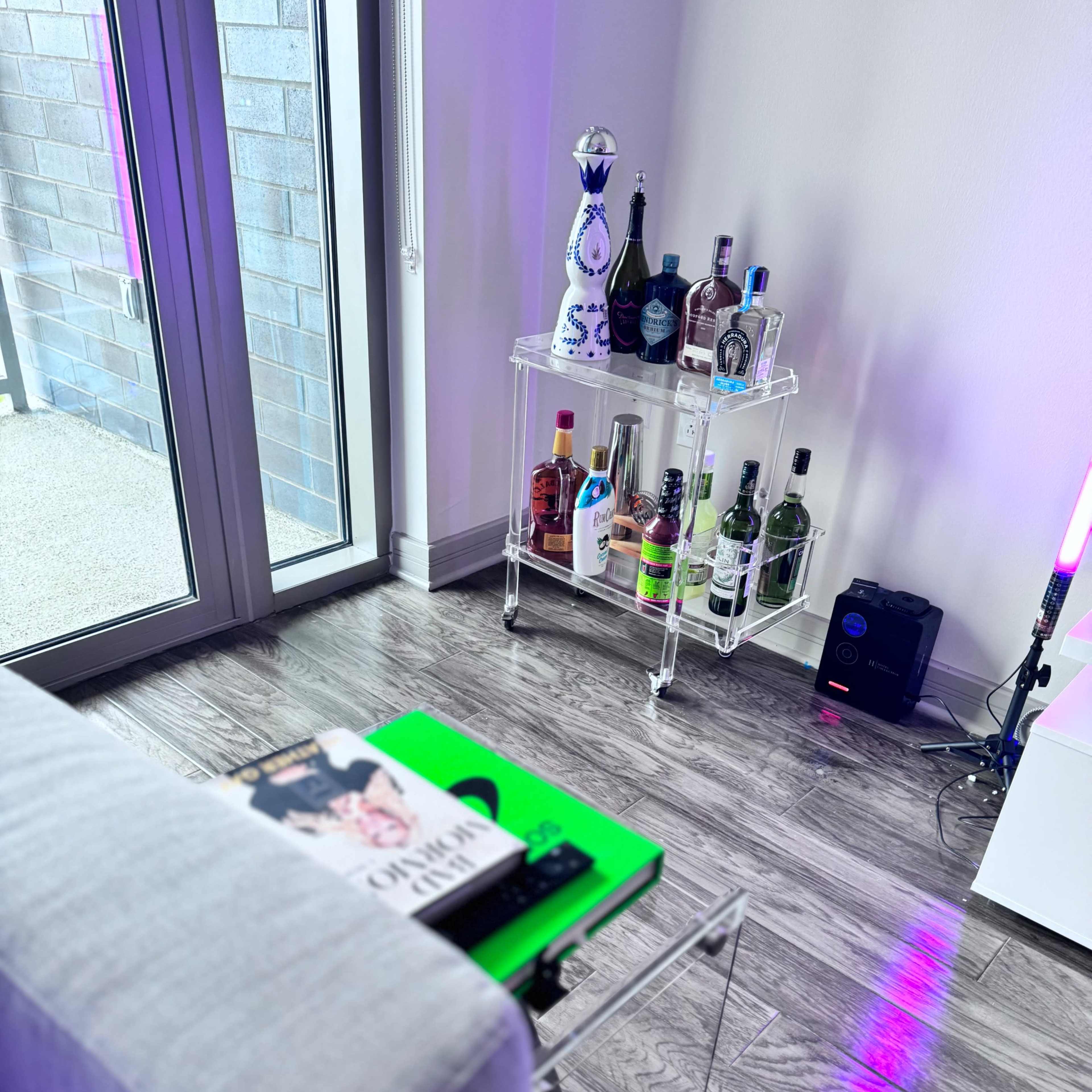 A modern living room features a gray sofa, a clear acrylic side table with books, and a glass bar cart stocked with various bottles.