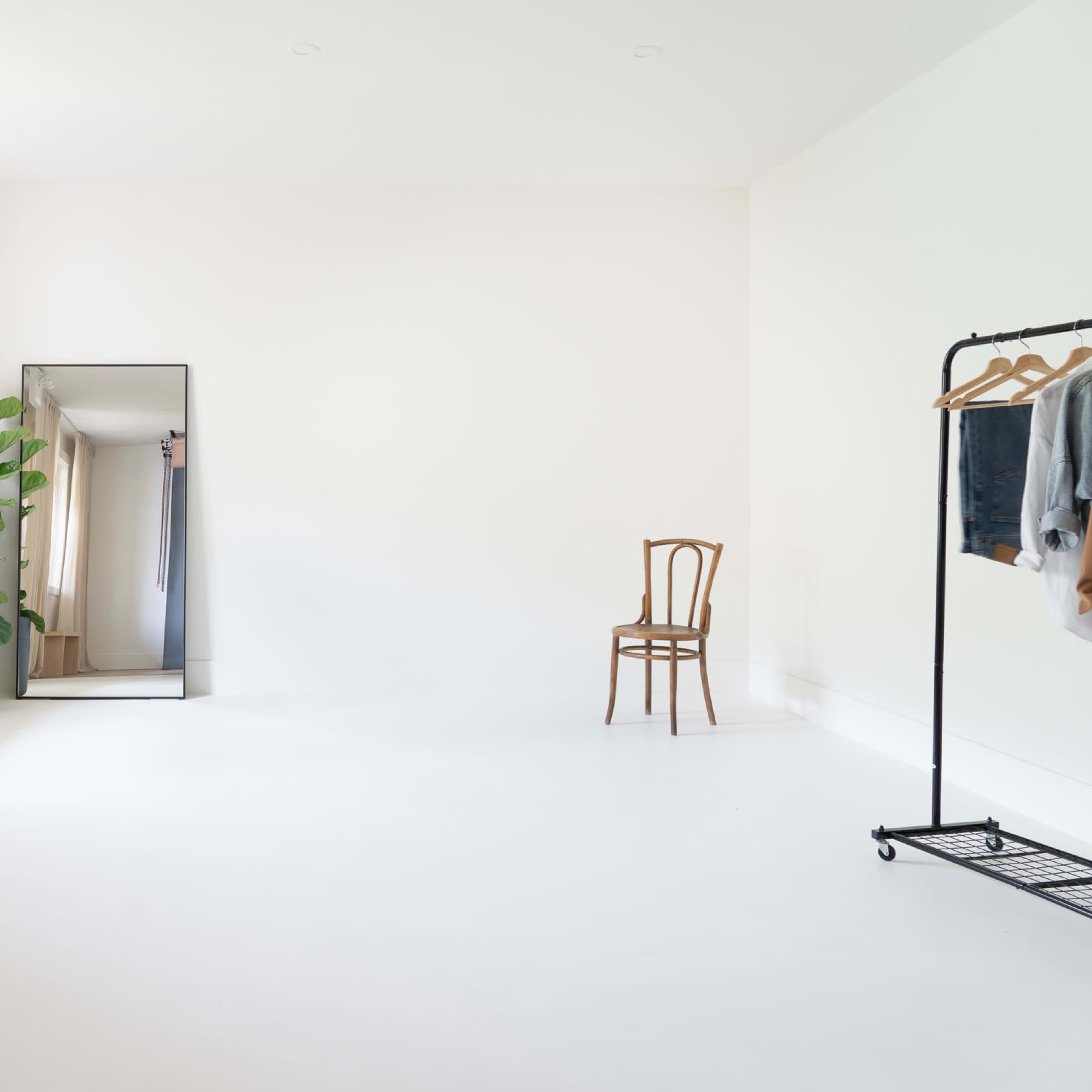 A bright, minimalist room features a tall mirror, a bench, a potted plant, a metal clothing rack with garments, and a wooden chair.