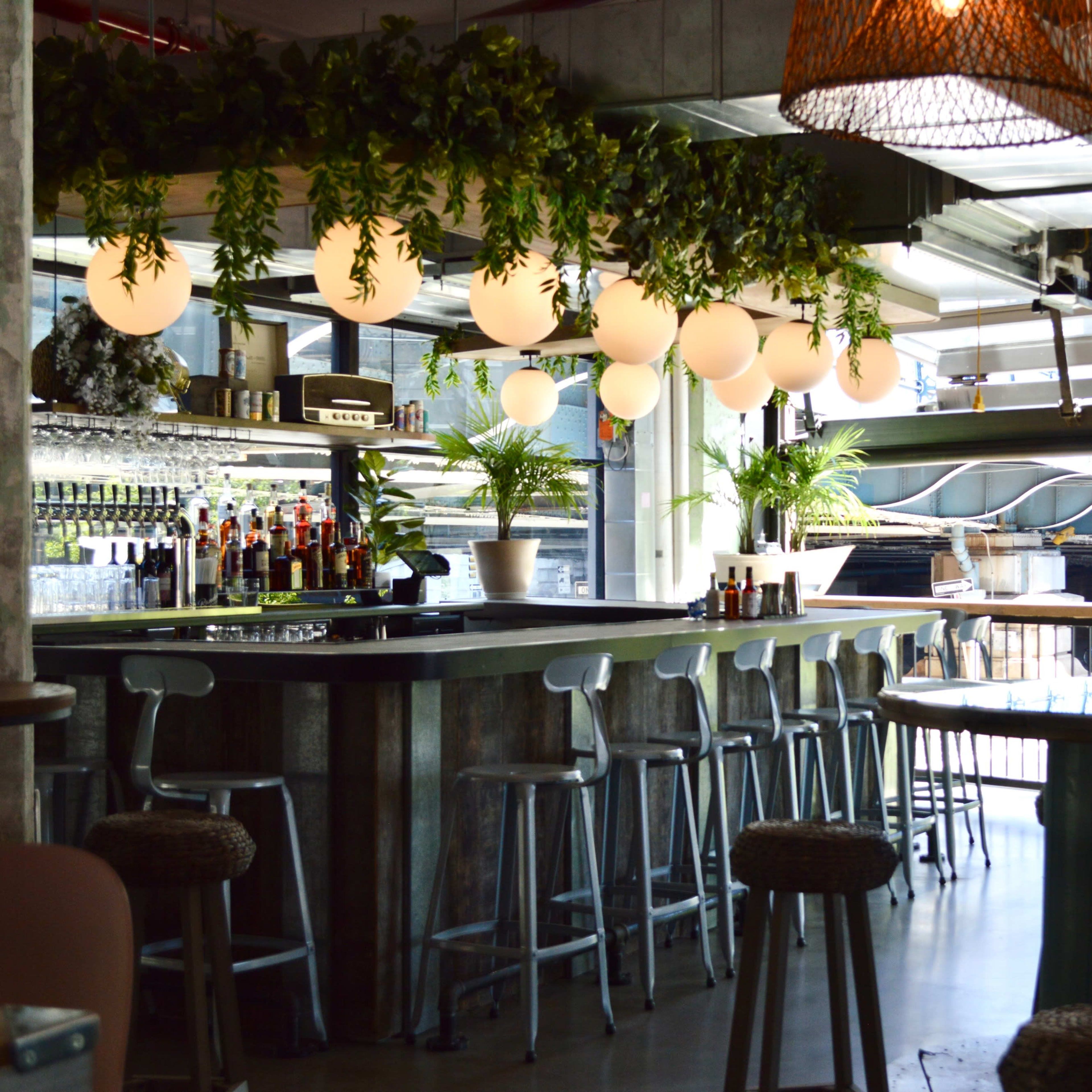 A modern bar interior with a long counter, pendant lights, and greenery decor.