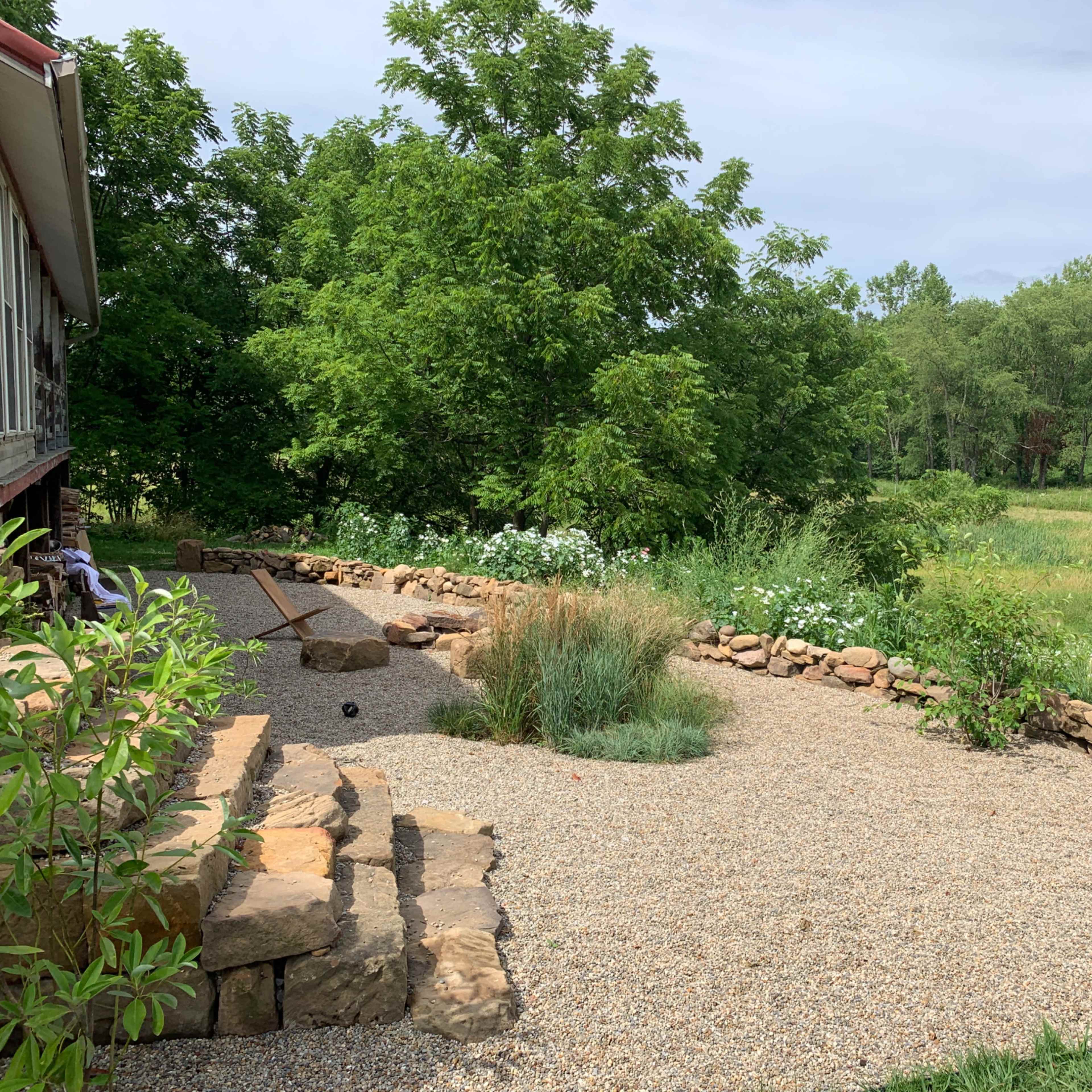 A landscaped area with gravel, stone edging, and plantings beside a wooden structure and a grassy field in the background.