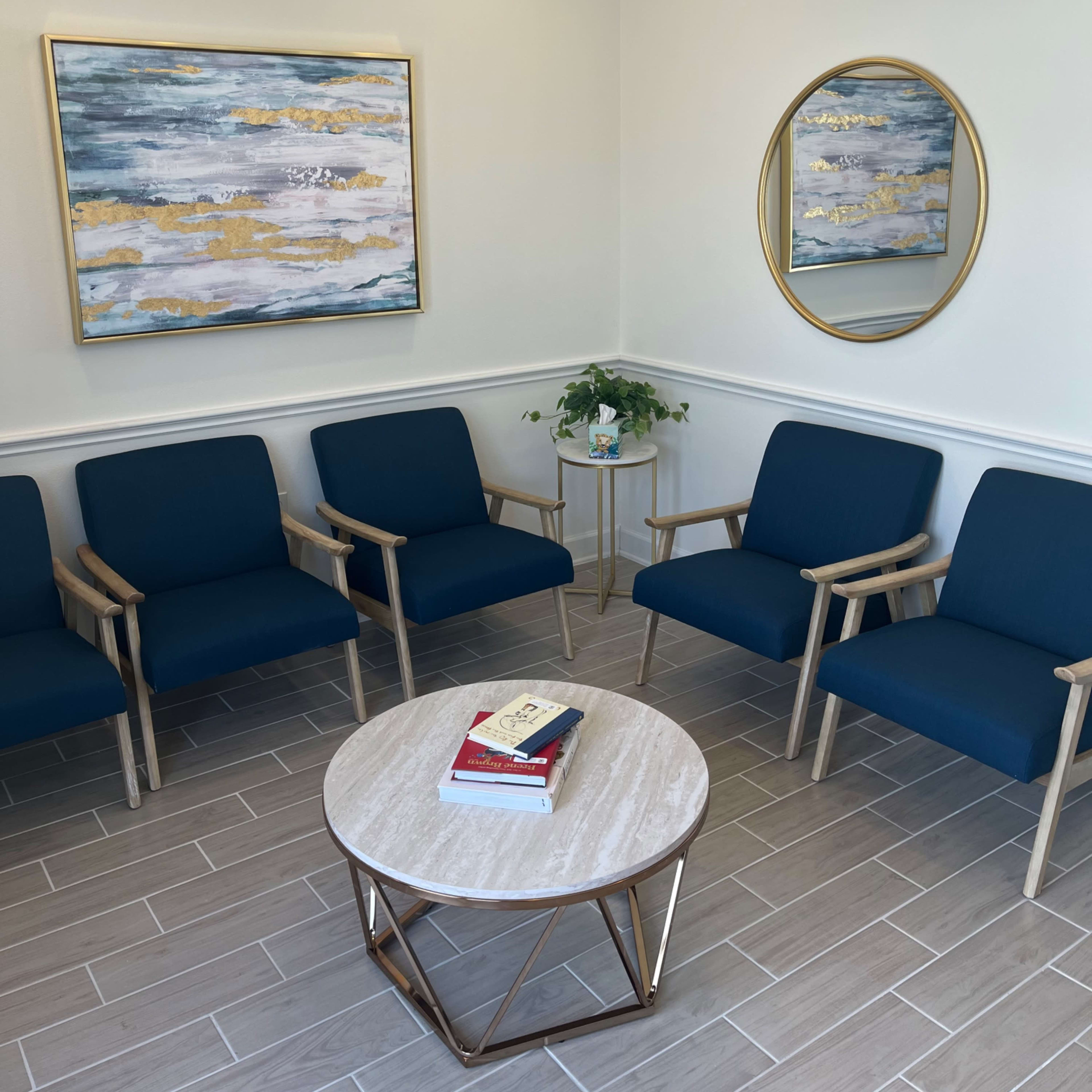 A waiting area with four blue chairs arranged around a round marble-top table, featuring two abstract paintings and a mirror on the walls.