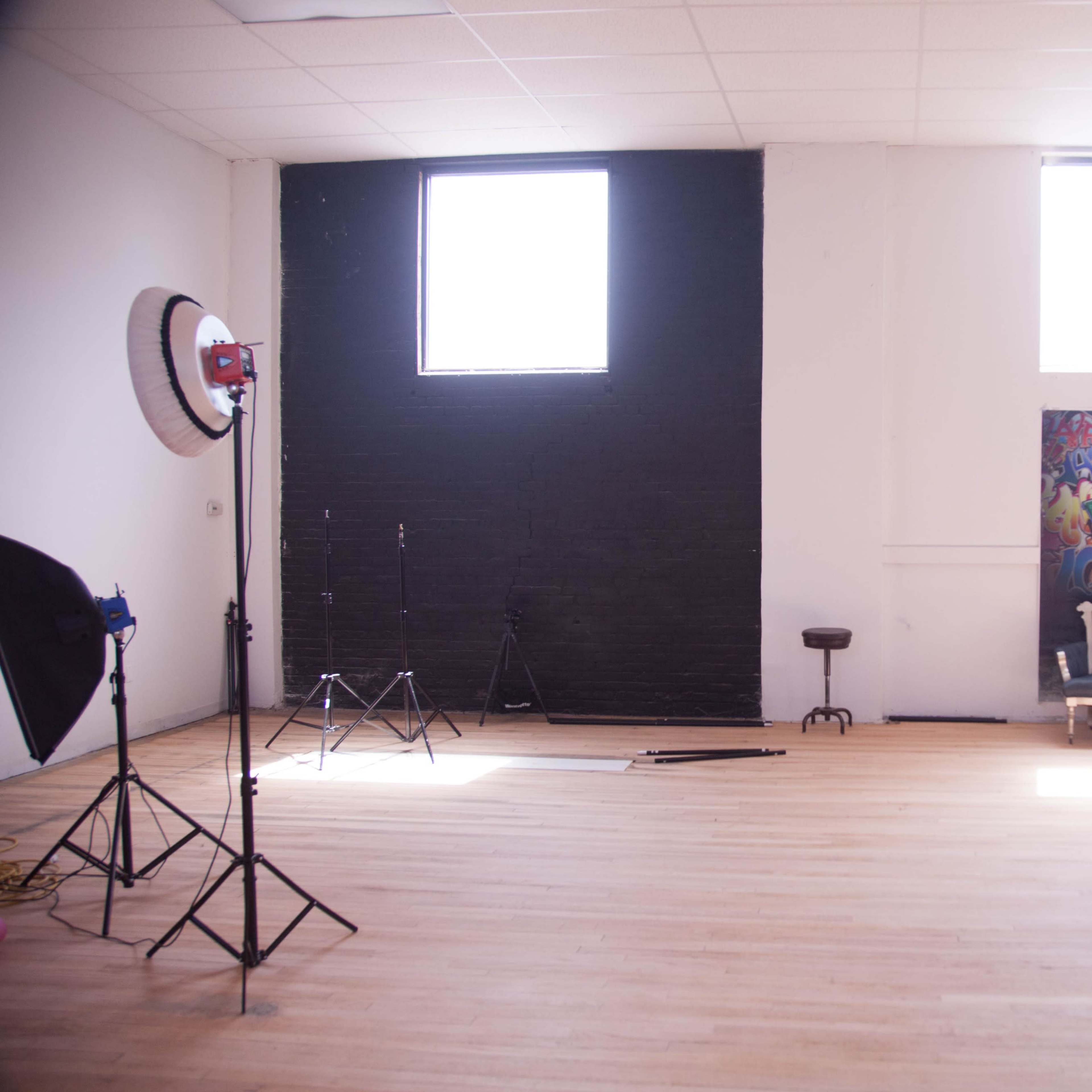 A spacious room with hardwood floors, two windows, a black wall, a colorful graffiti mural, and a photography setup including a light stand and a stool.